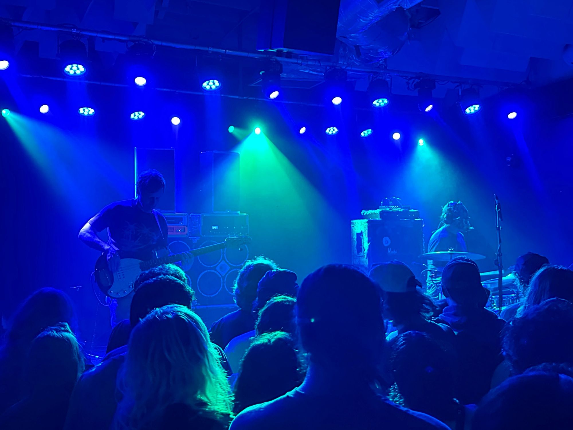 A man playing bass while another man wearing a mask is playing the drums. The back of the crowds head is visible in front. Everything is blue from the stage lights, and the band is heavily backlit.