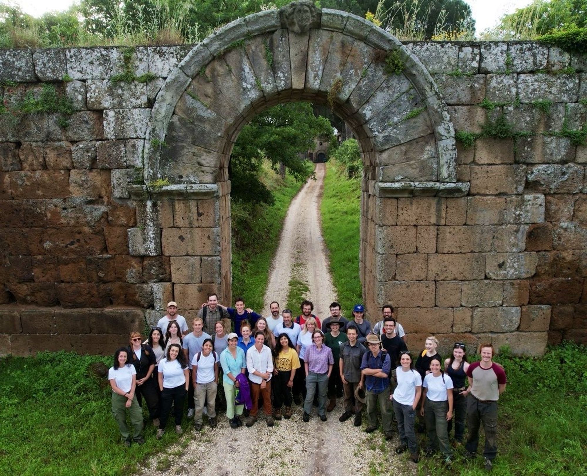 A group of people standing in front of an ancient Roman city gate and walls. A road continues behind to a farm and church.