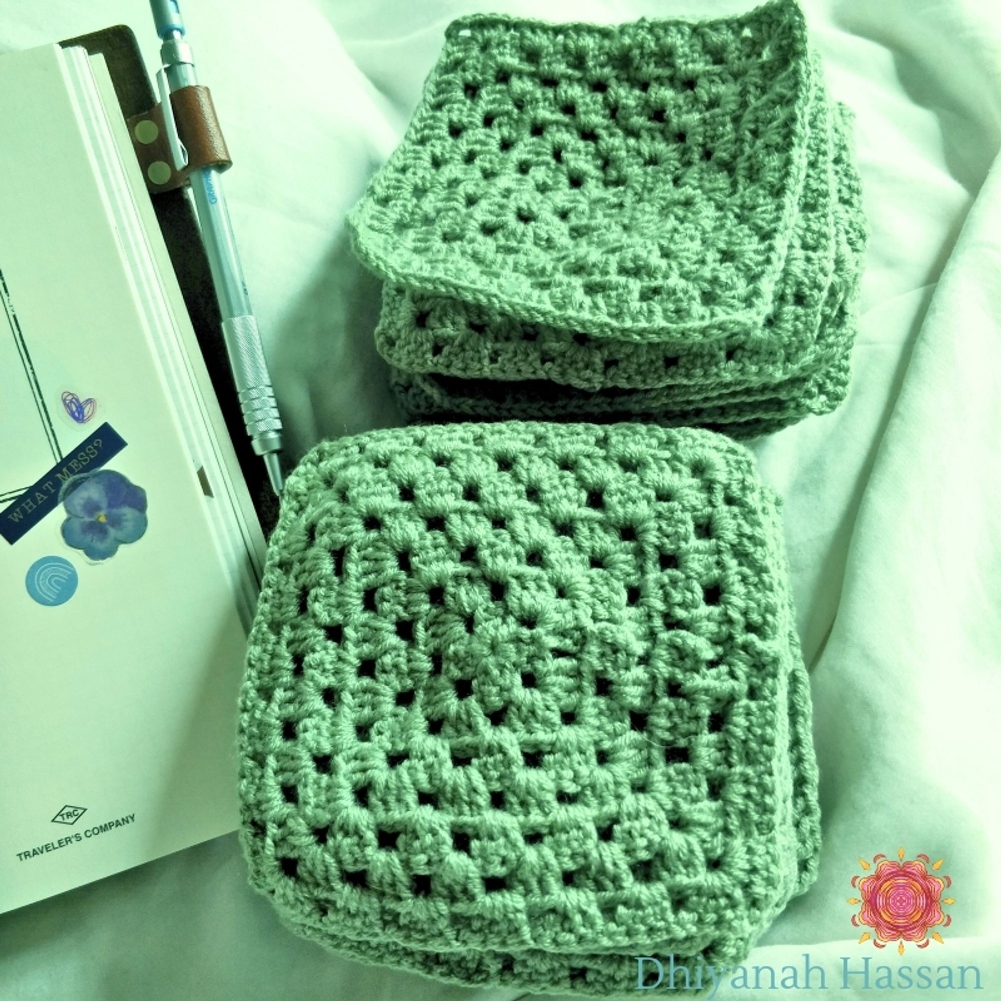 A photo of matcha green crochet granny squares stacked in two piles against a soft cream background. Beside them is my sketchbook with a mechanical pencil attached to it on the side.