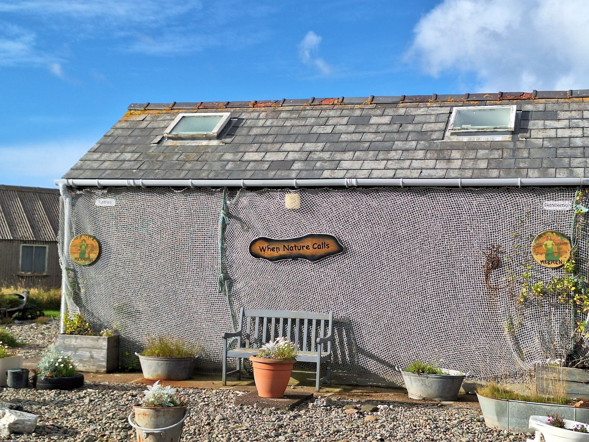 A public toilet in Hillswick. It's decorated with a fishing net. In front, there's a bench and flower pots. A sign on the wall says "When Nature Calls" and additional signs indicate "Mermaids" and "Mermen".