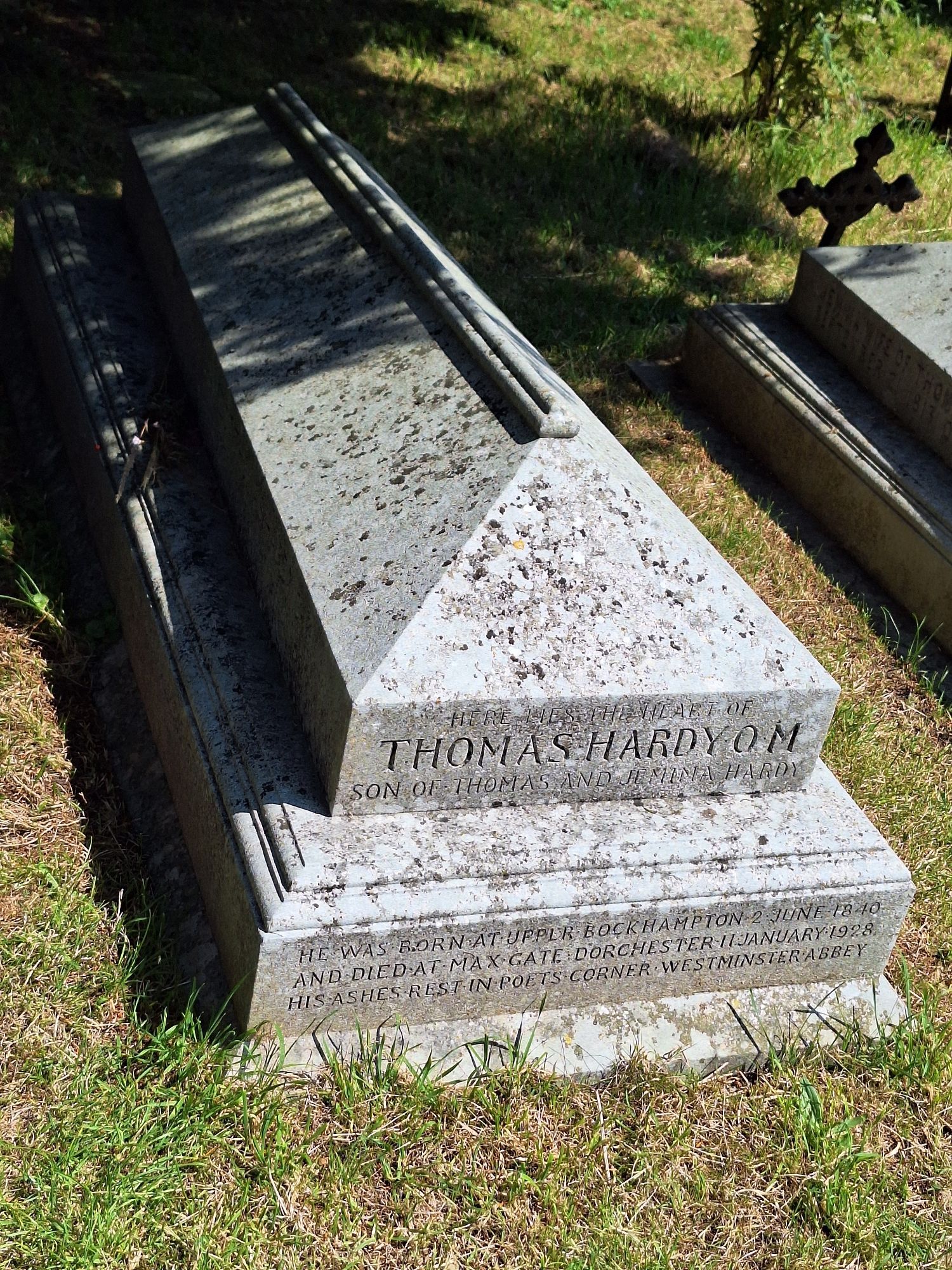 The stone grave of Thomas Hardy, next to it are other graves and you can see the grass surrounding it. On the front of the grave,  it says: 
"HERE LIES THE HEART OF THOMAS HARDY OM
SON OF THOMAS AND JEMIMA HARDY 
HE WAS BORN AT UPPER BOCKHAMPTON 2 JUNE 1840 AND DIED AT MAX GATE DORCHESTER 11 JANUARY 1928
HIS ASHES REST IN POET'S CORNER WESTMINSTER ABBEY