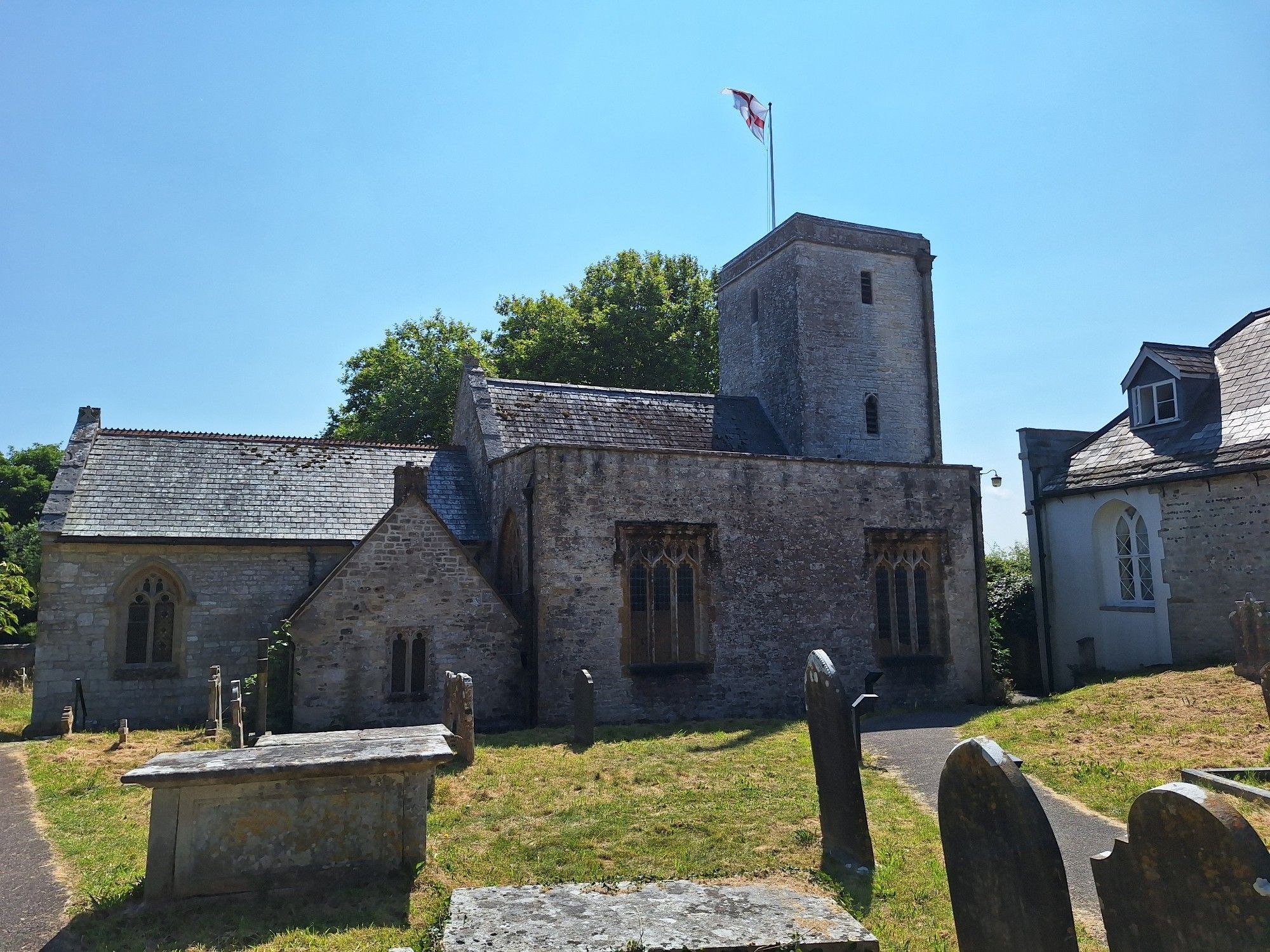 St. Michael, Stinsford. A church made of grey stone with an England flag on top of the church tower. In front, there's the graveyard witj various grave stones visible.