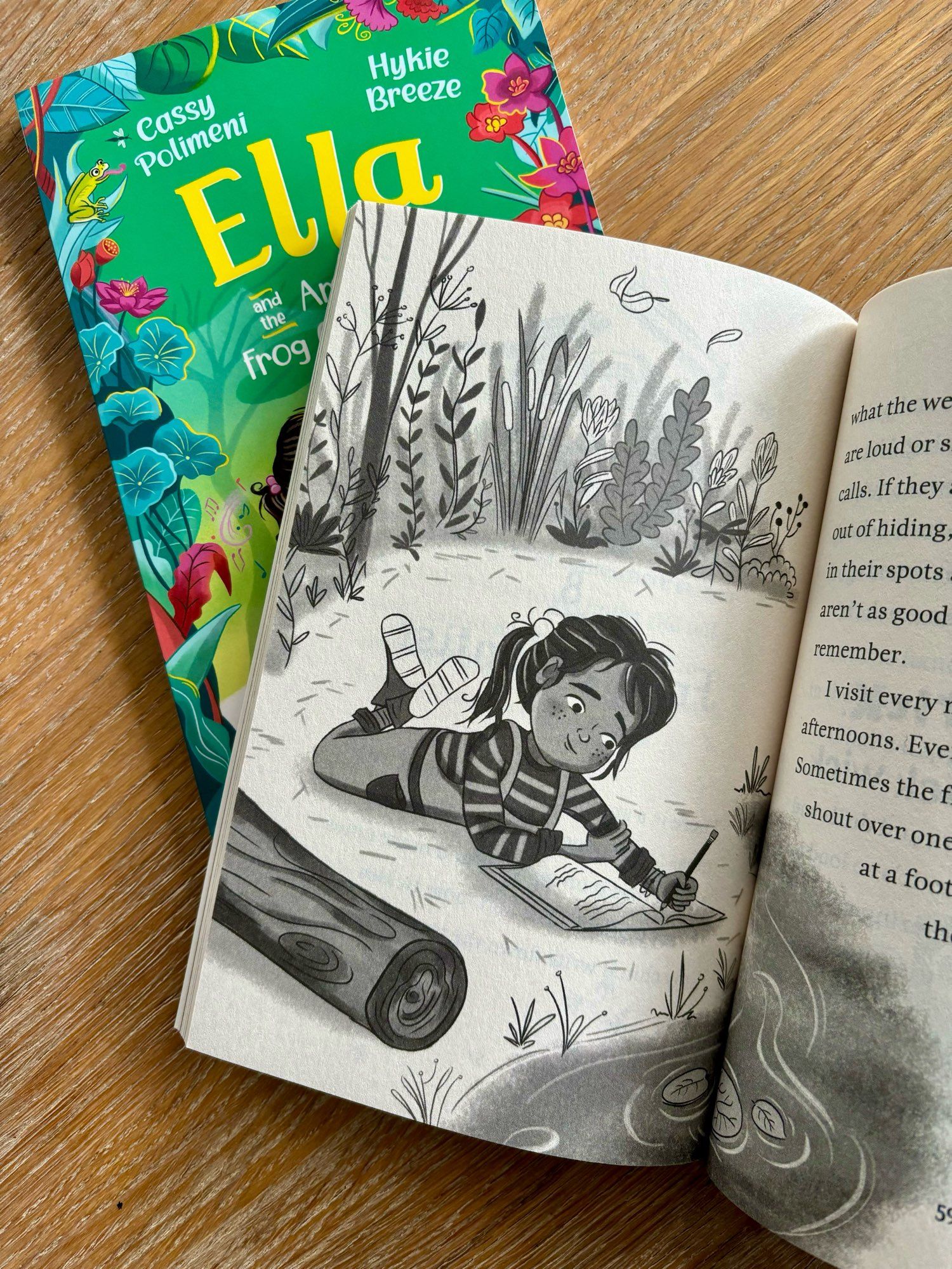 Inside the pages of Ella and the Amazing Frog Orchestra, a new middle grade fiction book. A black and white illustration of the main character, an 8 year old girl, lying outdoors by a frog pond, taking notes in her journal.