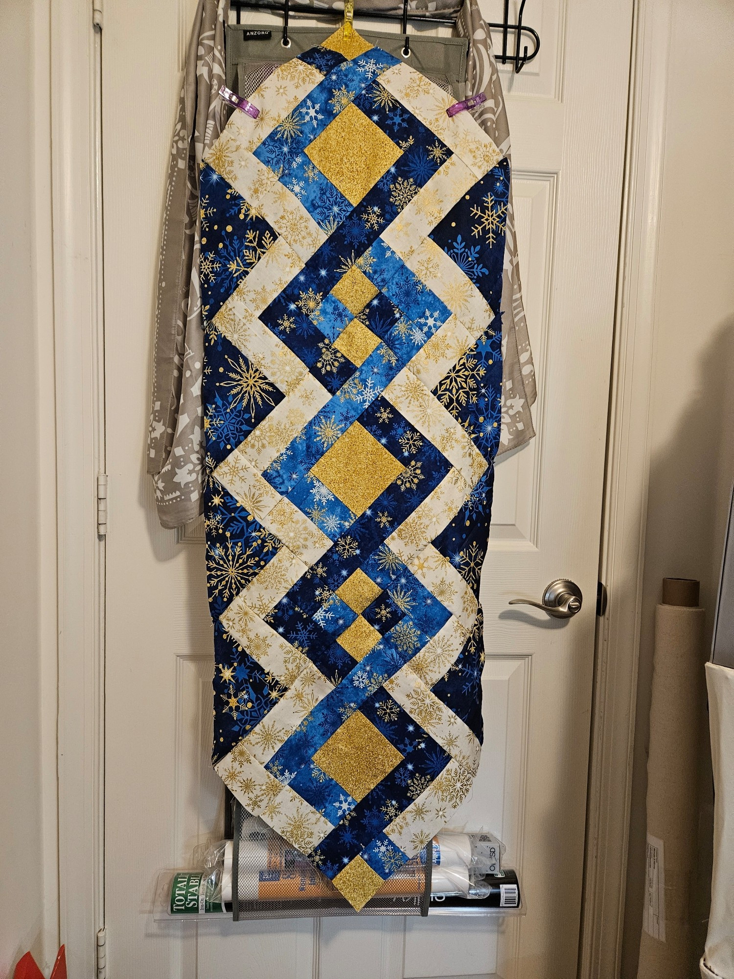 A lattice pieced quilt top for a holiday table runner made of alternating light blue fabric, dark blue fabric, cream fabric - each covered with coordinating snowflakes - and mottled gold fabric, and all the blocks set on point.