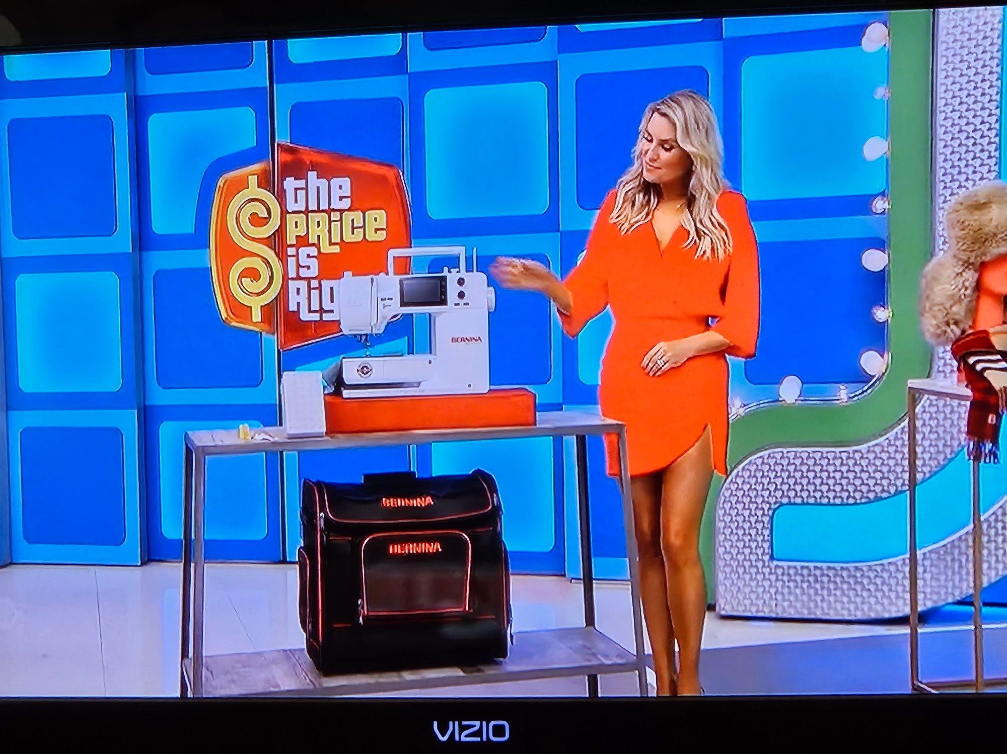 The Price Is Right showing a white Bernina 475 sewing machine and black trolley bag with a blond lady in a short orange long sleeved wrap dress pointing at them.