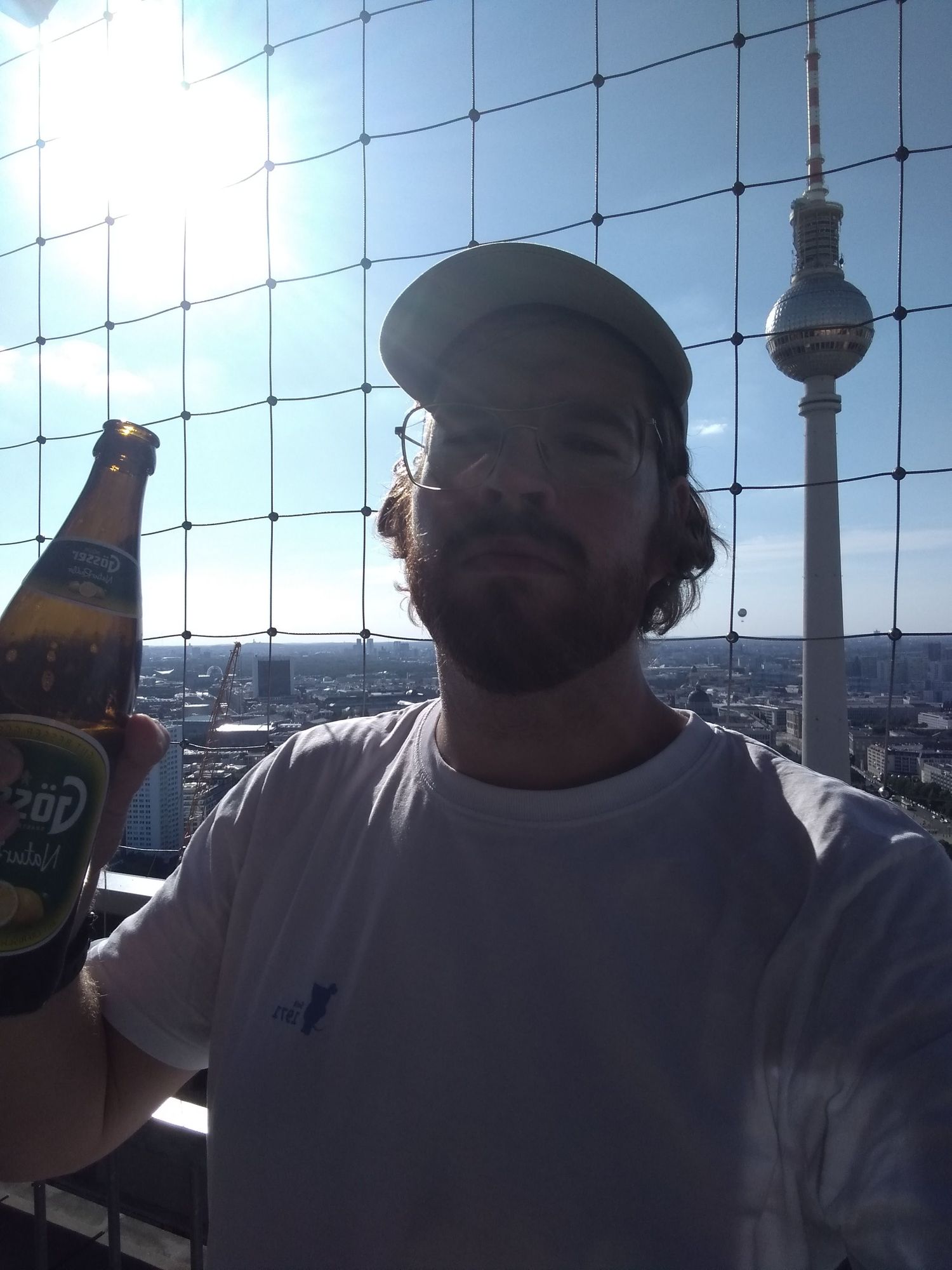 Selfie von mir, wie ich auf der Dachterrasse eines Hochhauses eine Flasche Gösser hochhalte. Im Hintergrund sieht man die Stadt Berlin mit dem Fernsehturm.