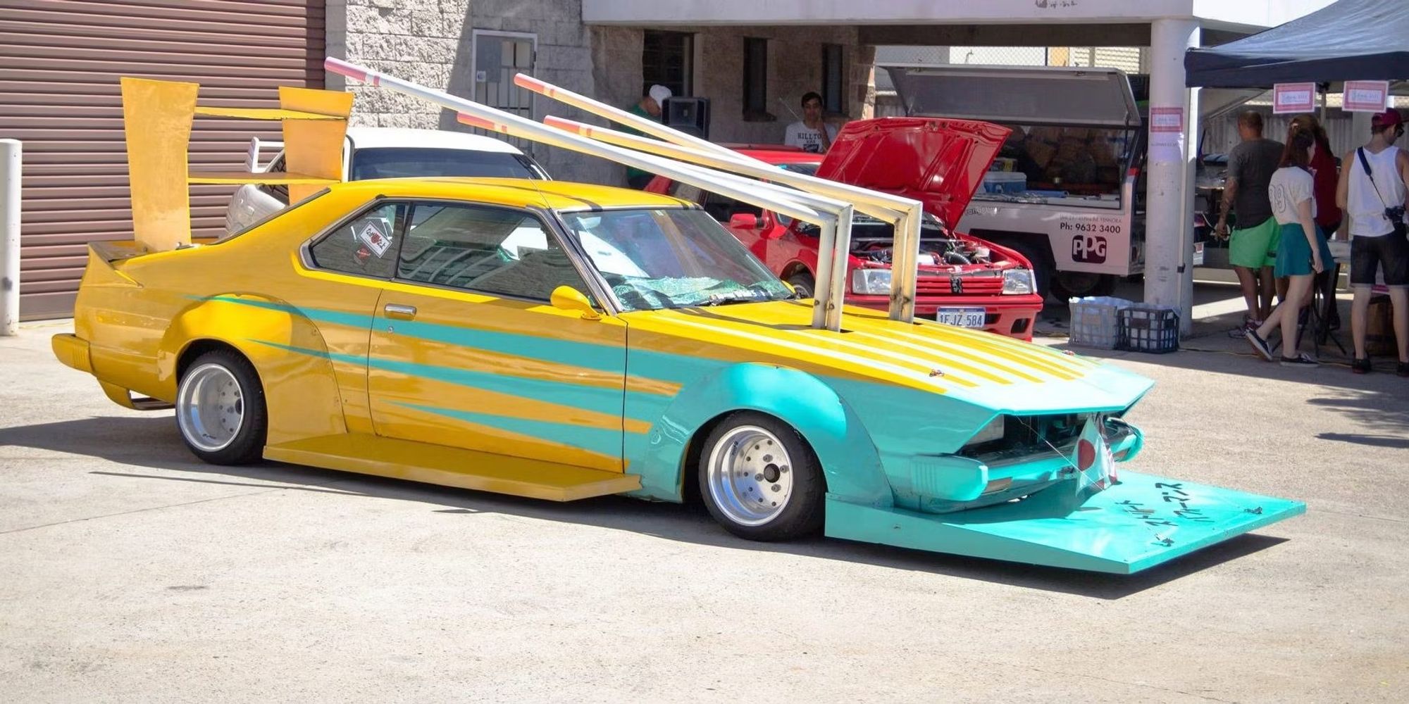 a teal and yellow lowered bosozoku-styled car with elongated underbite front scoop, towering double finned spoiler and hood exhaust pipes that extend far beyond the roof to the rear