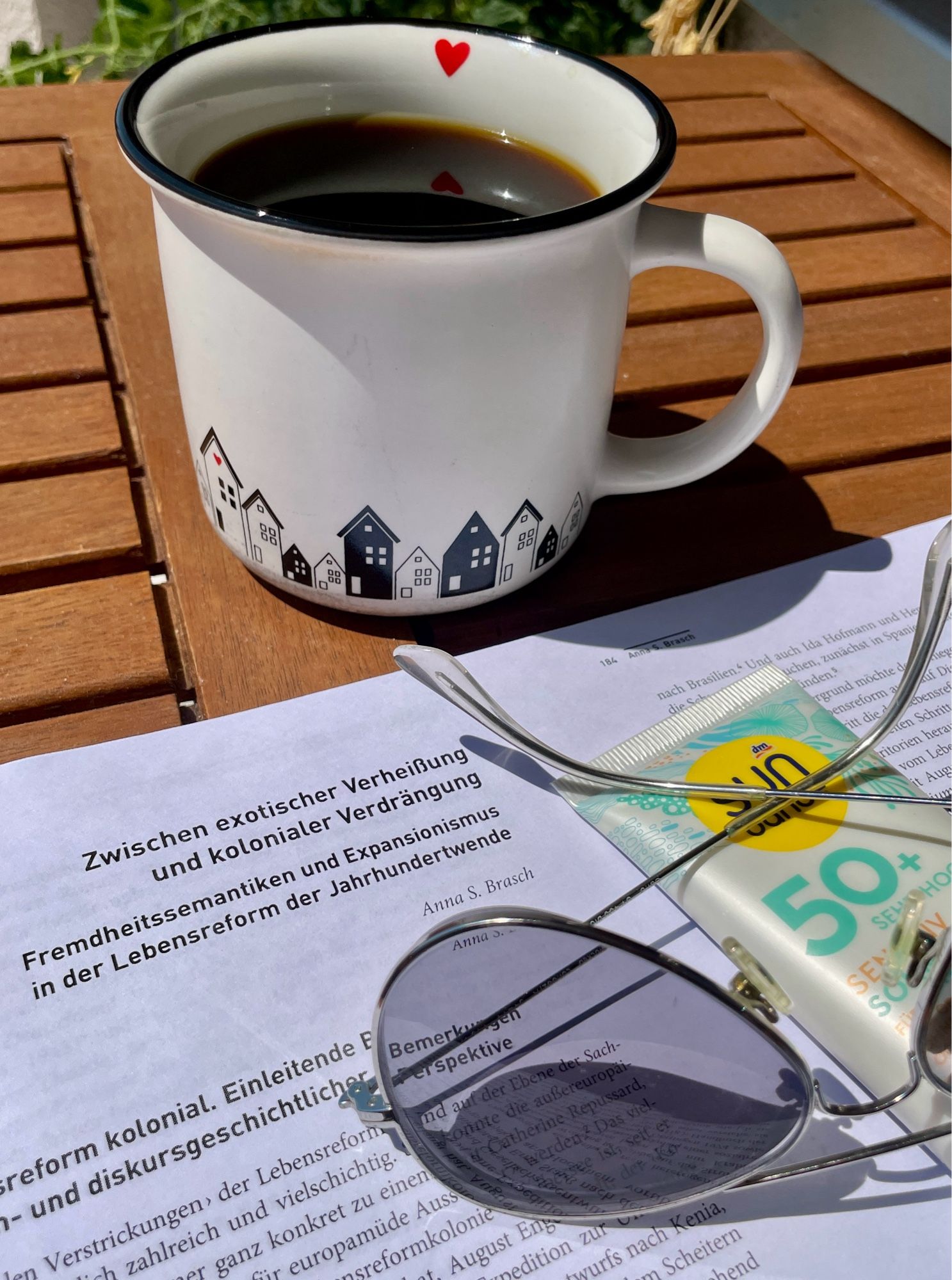 Druckfahnen, Kaffeebecher, Sonnencreme und Sonnenbrille auf einem Balkontisch