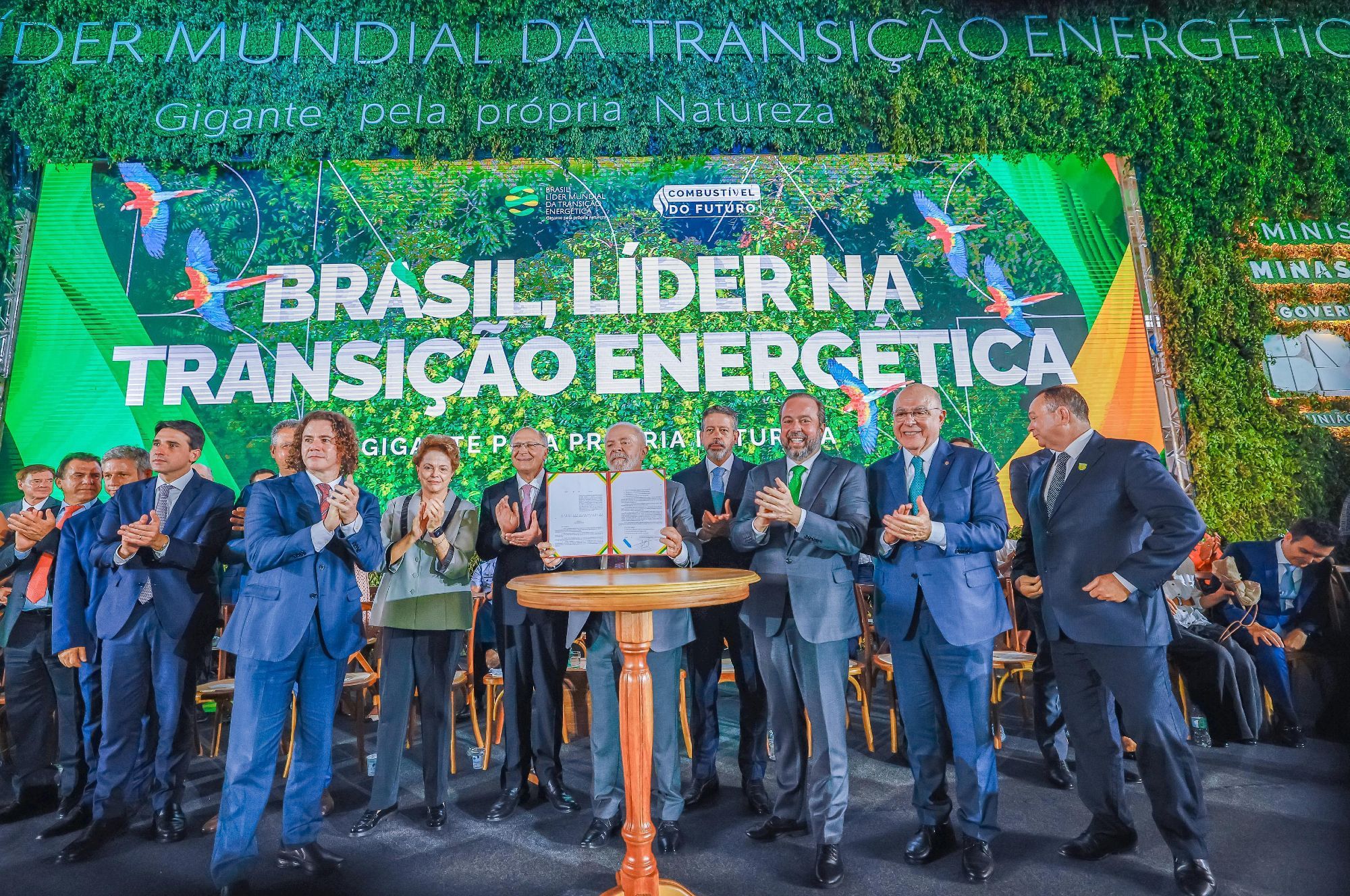 Na foto, presidente Lula, a ex-presidenta Dilma Rousseff e ministros estão no palco, segurando um documento assinado do programa Combustível do Futuro. Ao fundo, um texto no telão "Brasil líder na transição energética".