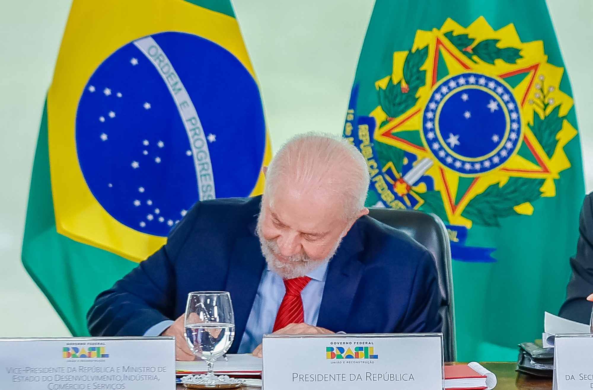 Na foto, o presidente Lula está sancionando um projeto de lei. Ele está assinando um documento em uma mesa. Ao fundo, as bandeias do Brasil e da República. O presidente usa uma gravata vermelha, um terno azul escuro.