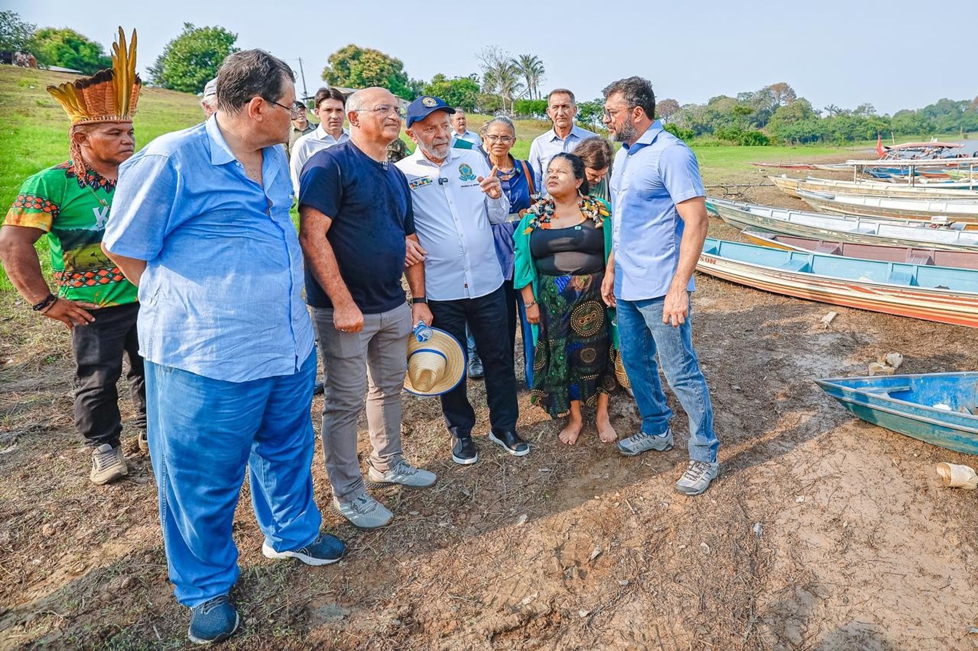 Em uma área aberta próxima a um rio, o presidente Lula, ao lado da comitiva de ministros, governador do Amazonas e prefeito de Manaquiri estão reunidos em frente a vários barcos atracados na margem. No centro da imagem, o presidente Lula, usando uma camisa branca com o brasão do Brasil e boné azul. Ao seu lado, está o governador do Amazonas, Wilson Lima, vestindo uma camisa azul clara, e outras autoridades, incluindo as ministra Sonia Guajajara e Marina Silva, uma liderança indígena com cocar, e outros ministros de governo. Todos estão em pé sobre o solo seco, que parece afetado pela estiagem, e ao fundo há árvores e um céu claro.