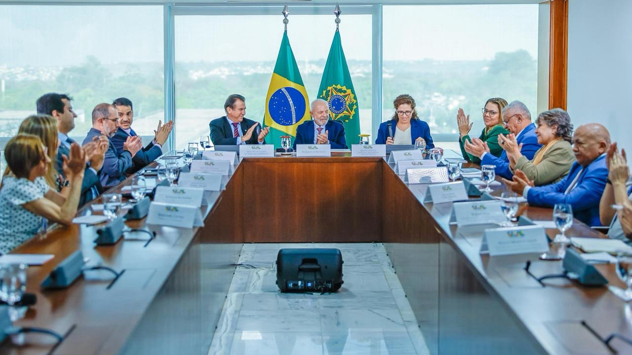 O presidente Lula ao lado de ministros e ministras, parlamentares e representantes dos trabalhadores terceirizados estão todos sentados à mesa e batem palmas. Ao fundo, as bandeiras do Brasil e da República.