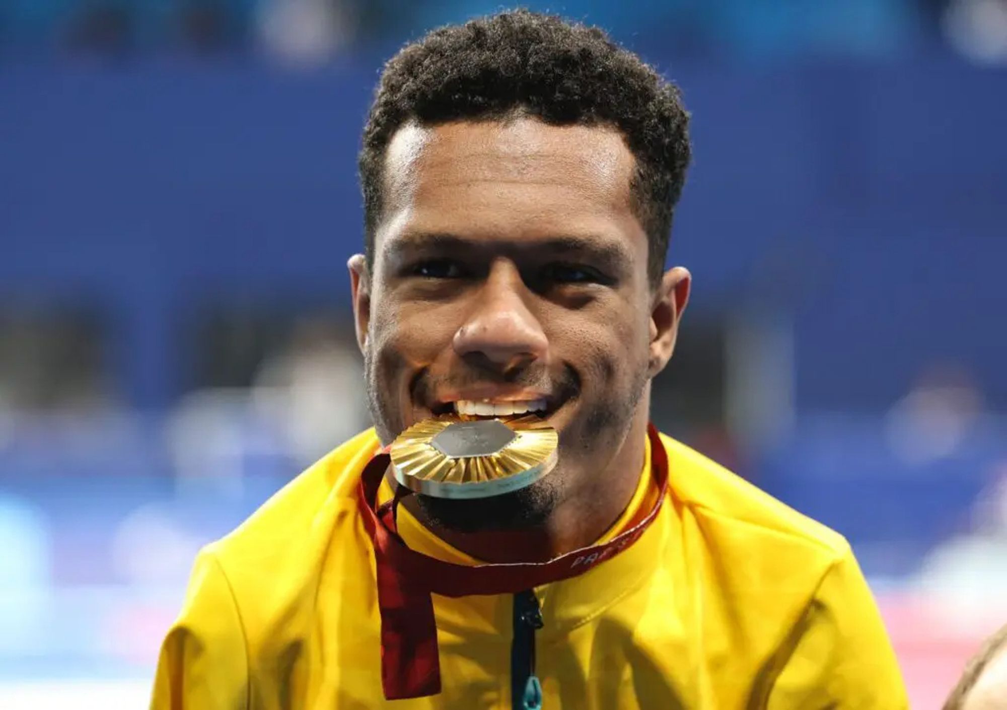 Na foto, o atleta paralímpico Gabriel Araújo com a sua medalha de ouro entre os dentes, sorrindo.