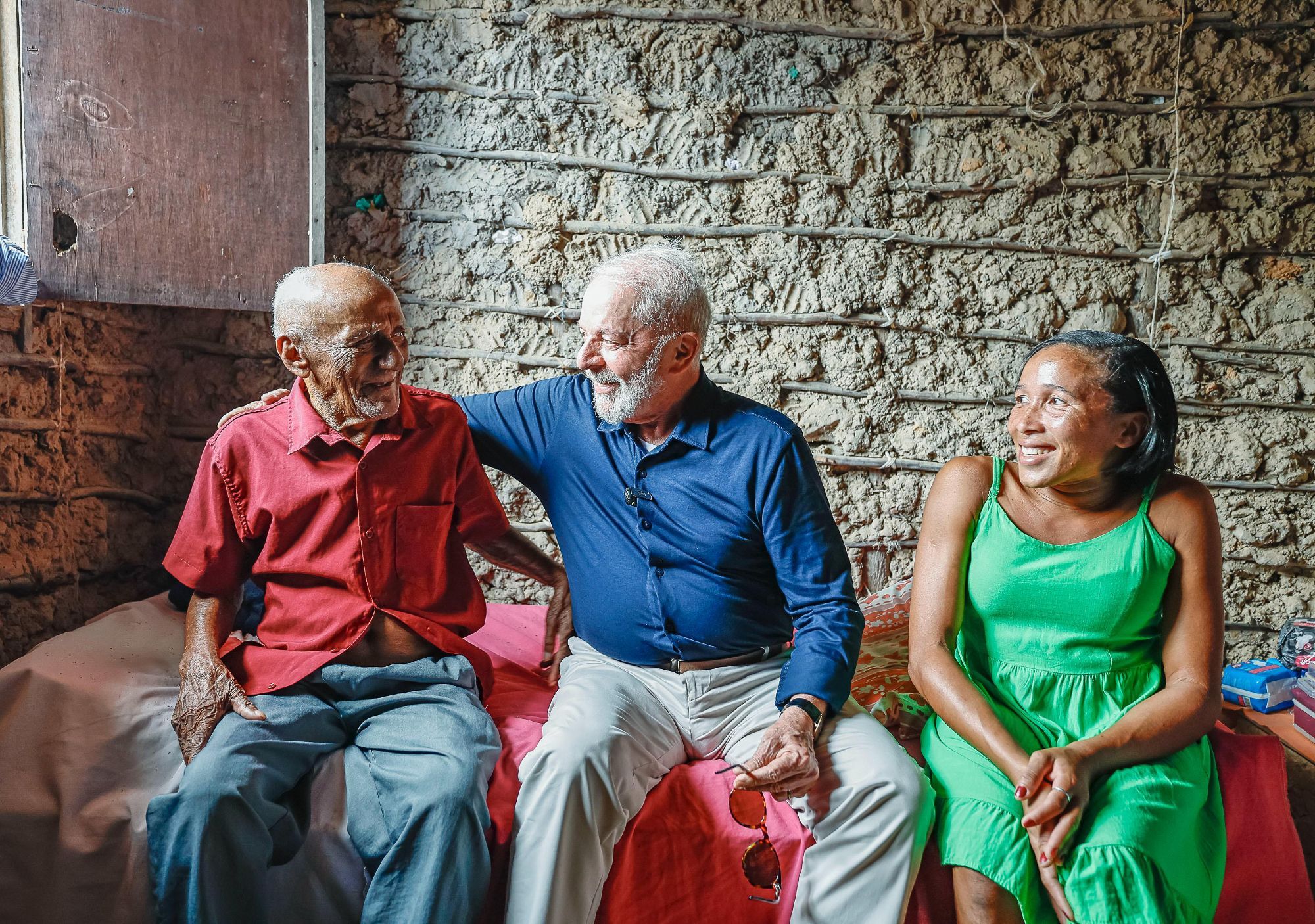 O presidente Lula ao lado dois moradores do Quilombo Mamuna, no Maranhão. Os três estão sentados na cama, conversando. Todos sorrindo.