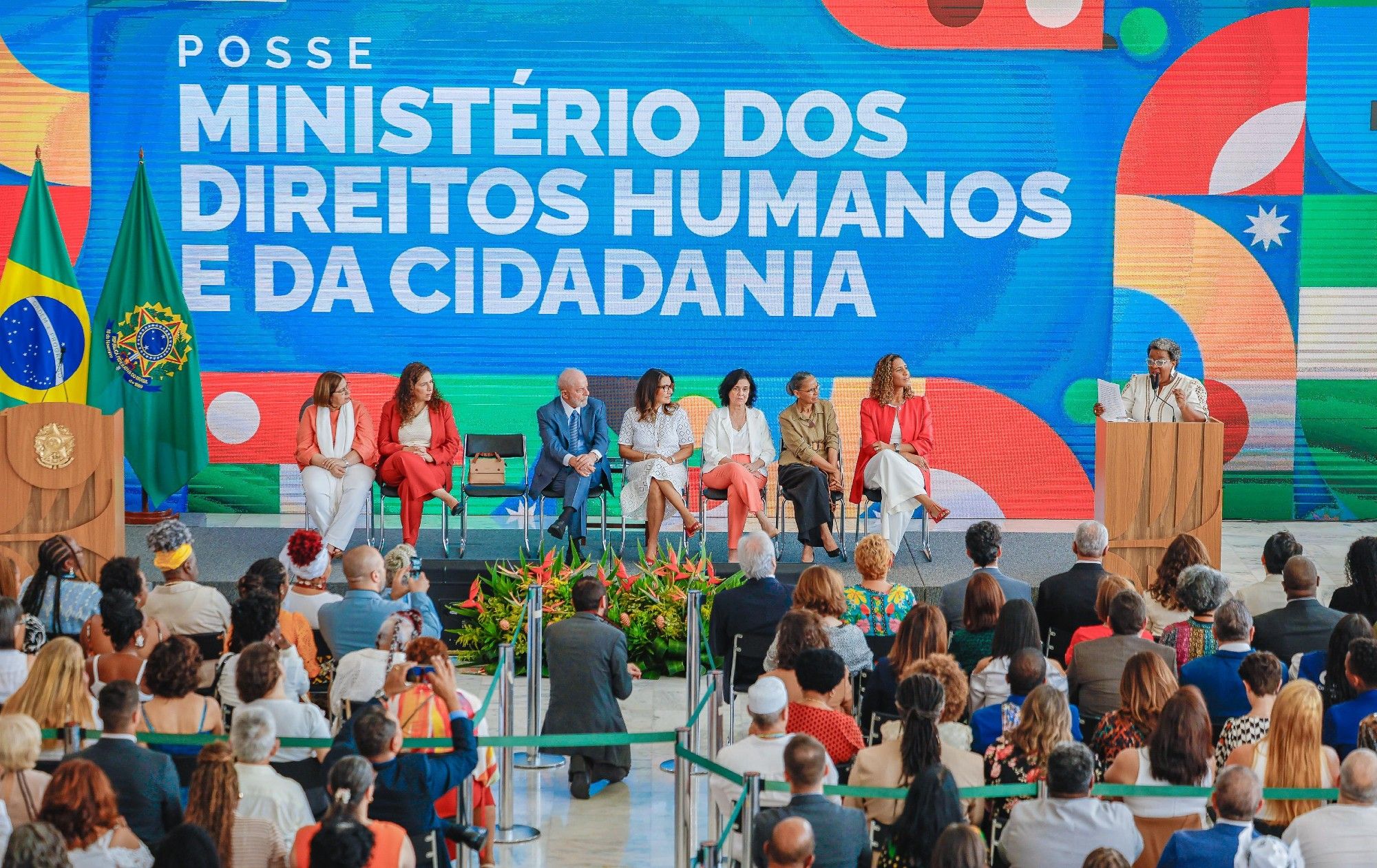 A foto mostra o presidente Lula, primeira-dama Janja, e as ministras Anielle Franco, Esther Dweck, Cida Gonçalves, Macaé Evaristo, Nísia Trindade e Marina Silva. Macaé está em pé, falando. Os demais sentados no palco. Ao fundo, o banner: posse Ministério dos Direitos Humanos e da Cidadania. O público assiste ao ato.