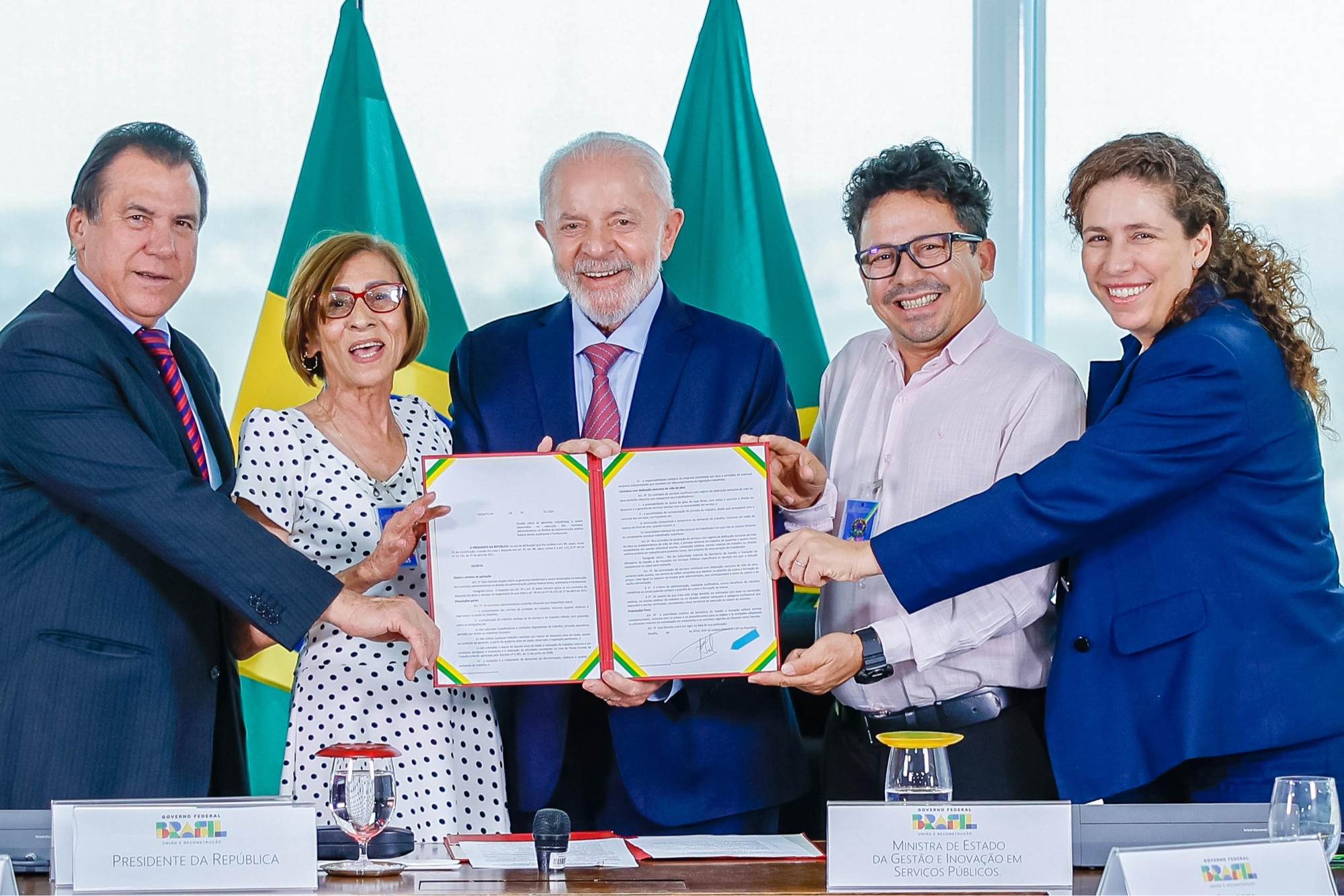 O presidente Lula, a ministra Esther Dweck e o ministro Luiz Marinho ao lado de dois representantes dos trabalhadores terceirizados seguram o decreto assinado pelo presidente. Todos sorriem para foto.