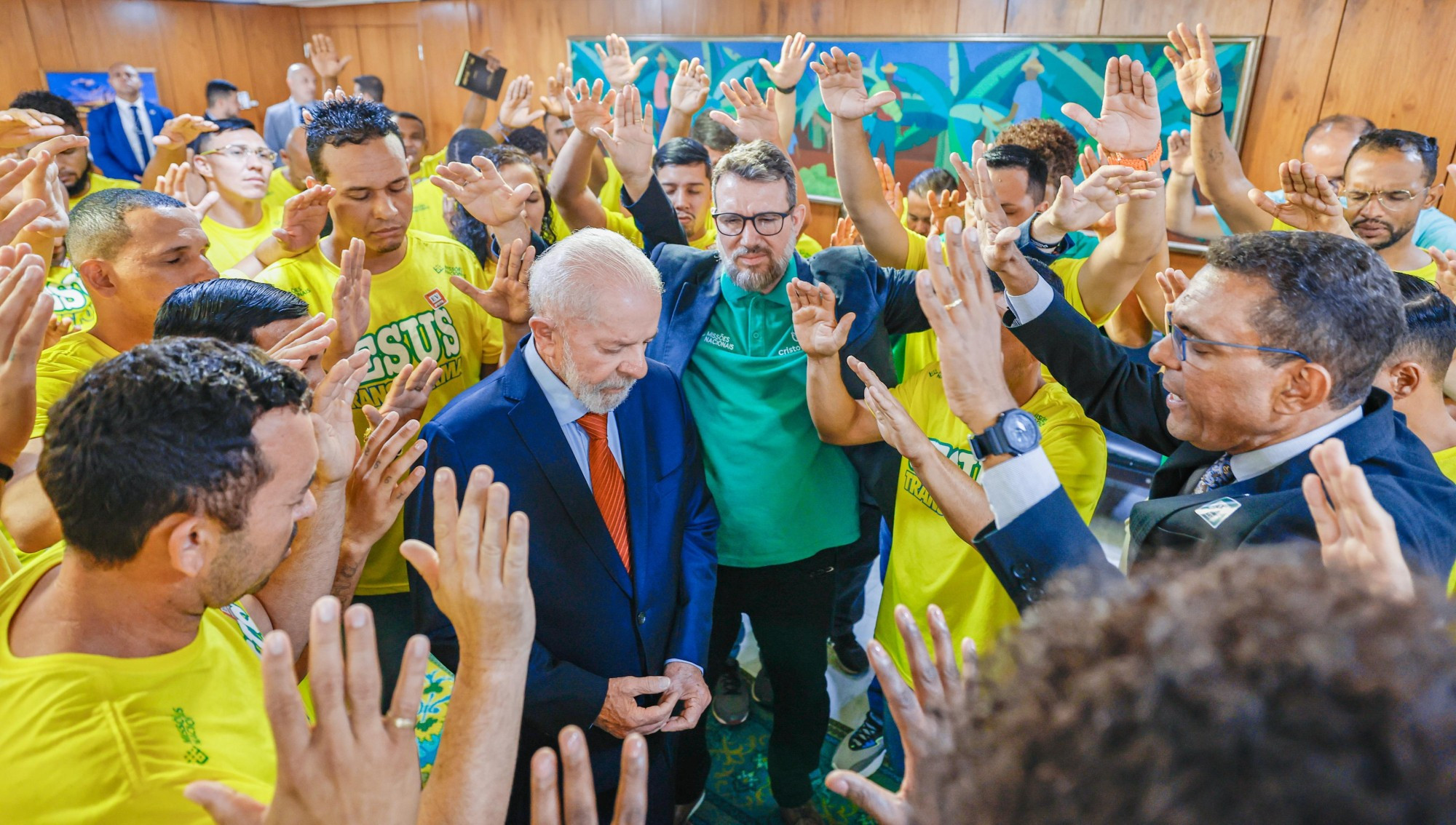 Na foto, o presidente Lula está de cabeça baixa, olhos fechados e recebendo as bençãos de representantes do Coral Cristolândia, que vestem camisas amarelas, e do deputado federal Otoni de Paula. 