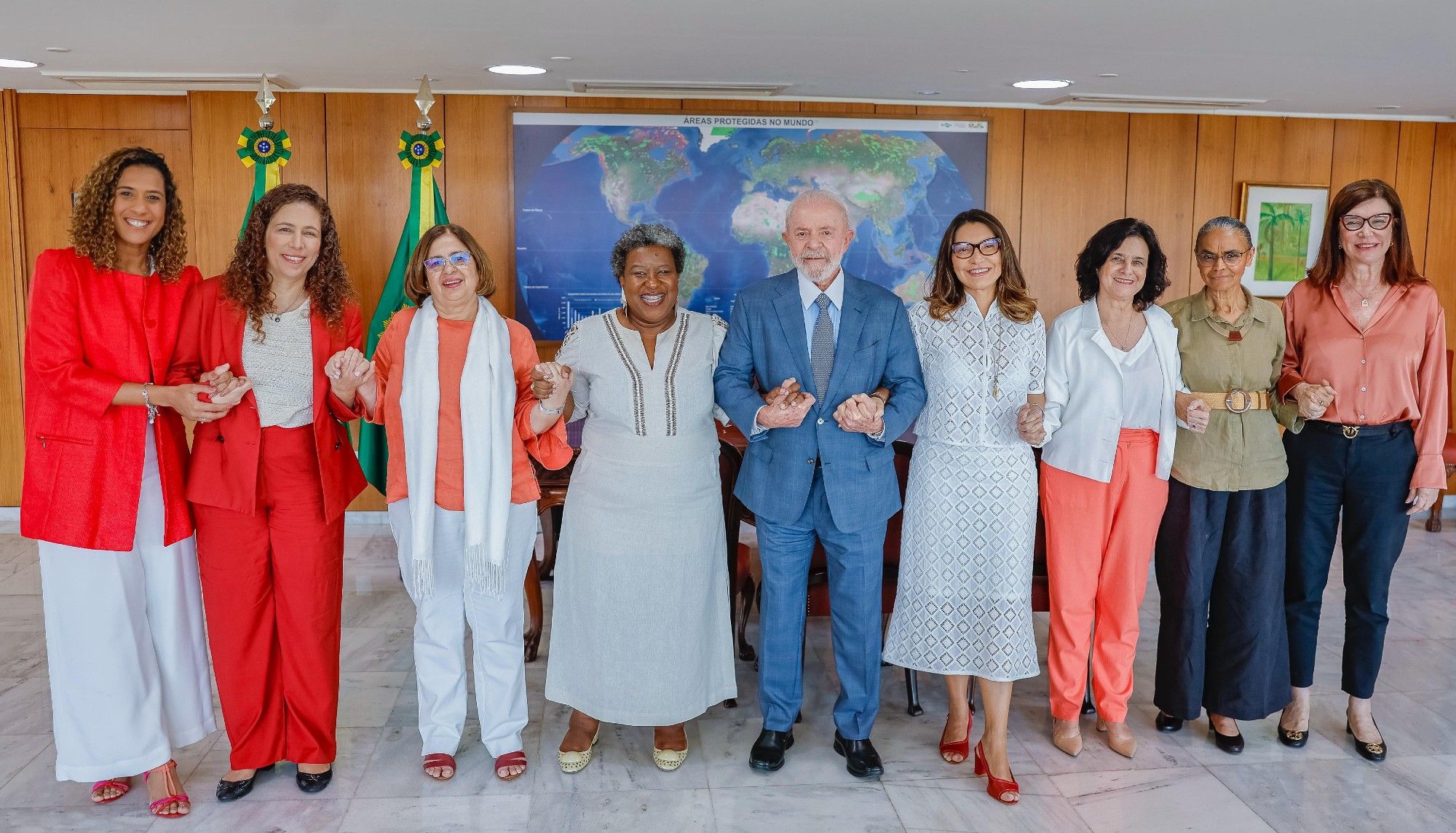 A imagem mostra o presidente Lula no centro, vestindo um terno azul claro, segurando as mãos de um grupo de ministras e da primeira-dama, Janja. Na ordem da esquerda para a direita: Anielle Franco, Esther Dweck, Cida Gonçalves, Macaé Evaristo, Lula, Janja, Nísia Trindade, Marina Silva e Magda Chambriard.