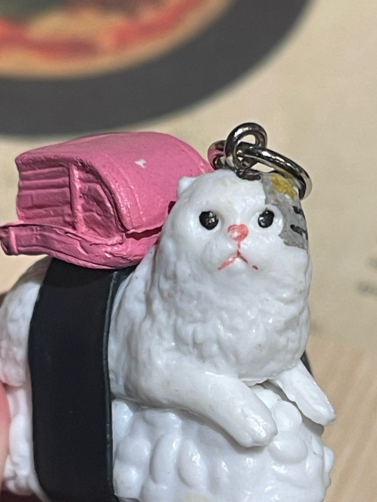 a close up shot of a cat sushi keychain. the cat is white with a gray patch, and has a little pink backpack at its back to imitate tuna. the cat stares stupidly into the void, without any thought at all.