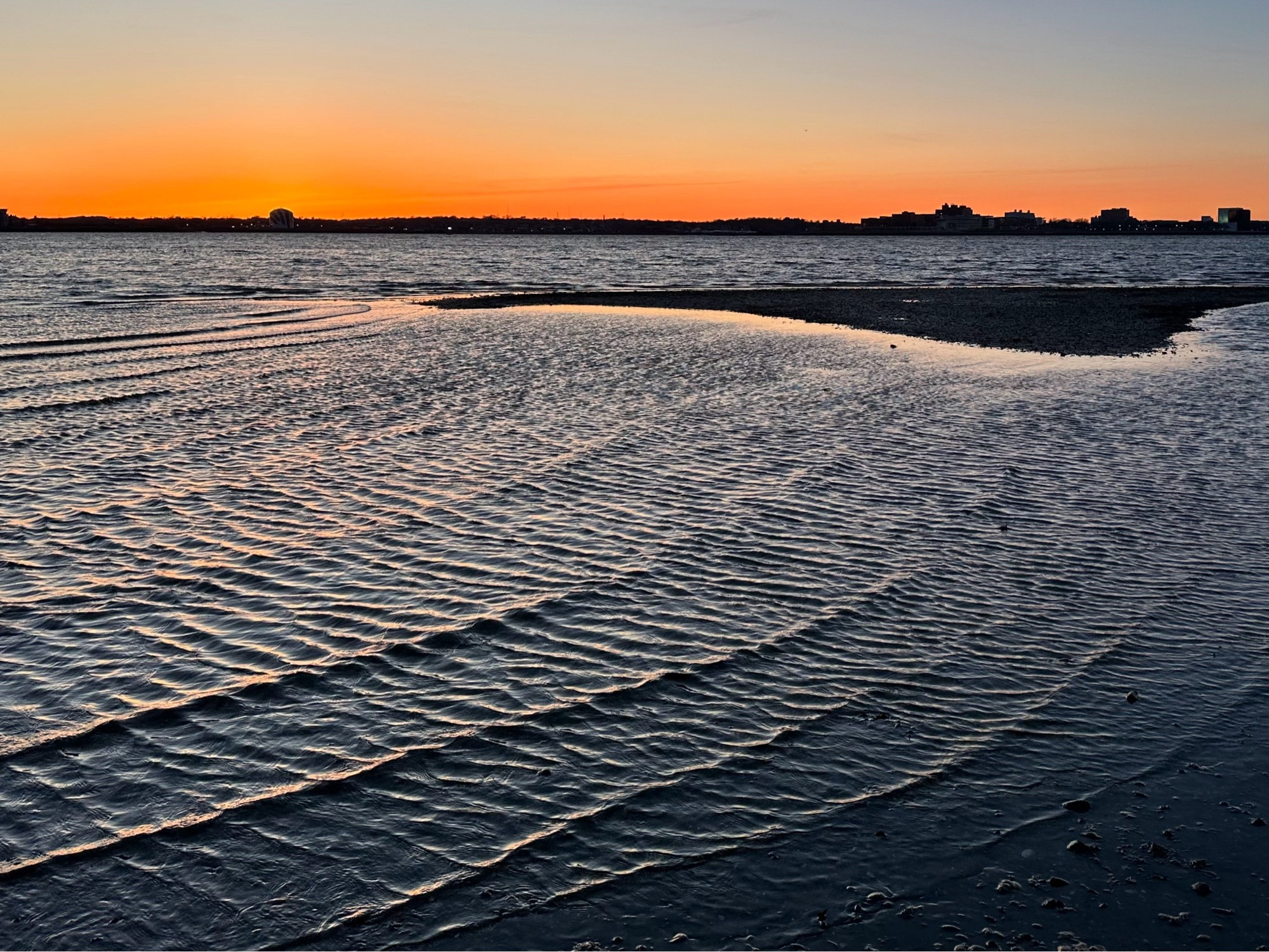 Twilight with orange sky and the combination of wind and a sandspit creating a bidirectional, orthogonally superimposed wave pattern in the ocean ripples.