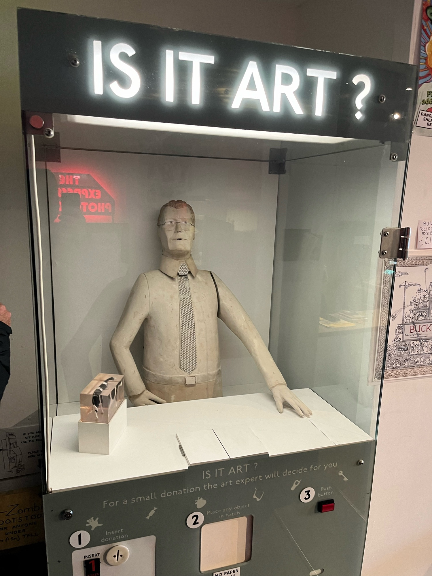 A photo of an arcade machine called is it art, where you insert objects and a the dummy of a man inspects it carefully and nods or shakes his head to indicate if it’s art