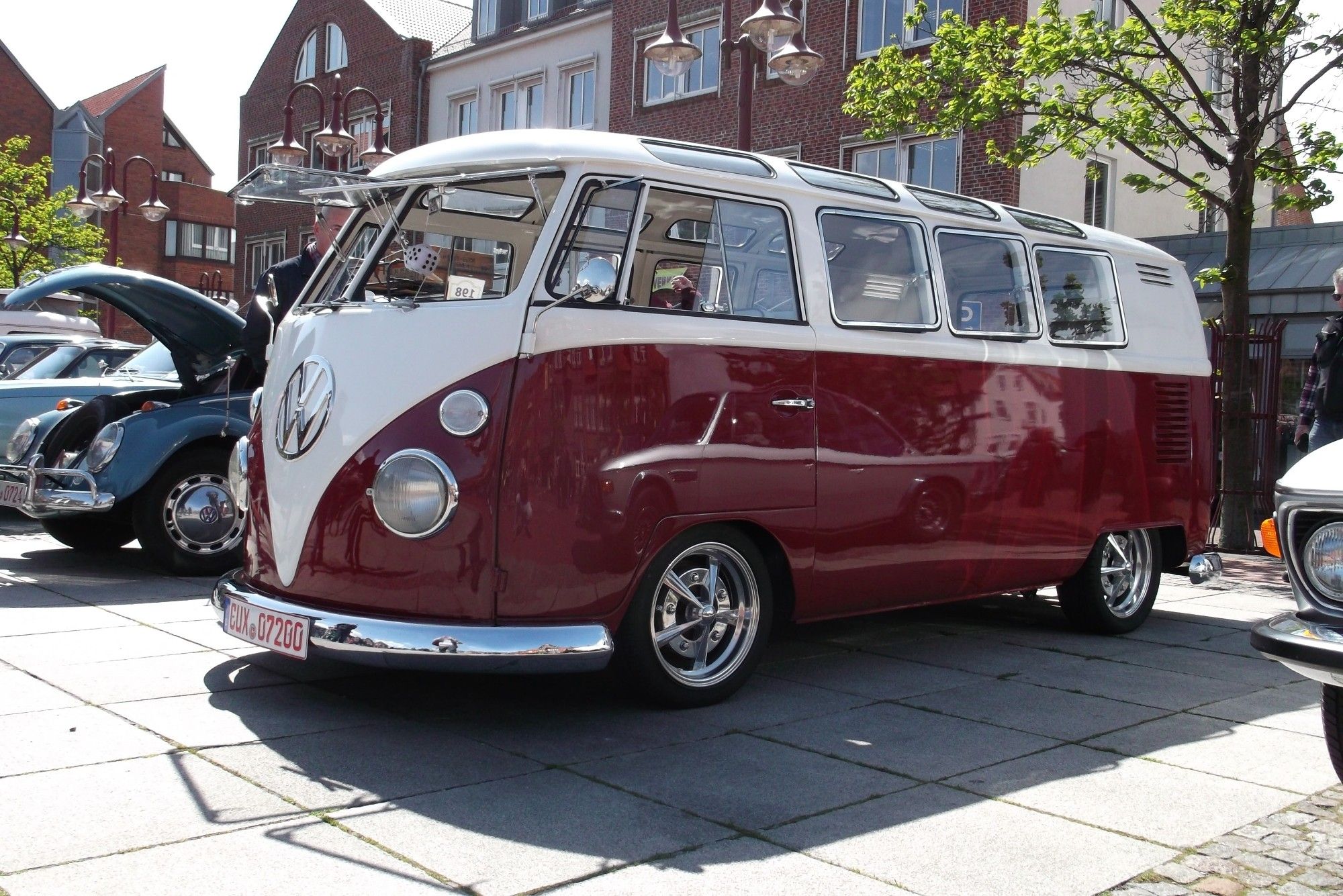 The image shows a classic Volkswagen T1 van, often referred to as a "VW Bus" or "VW Camper." The vehicle has a two-tone paint job, featuring white on the top half and a deep maroon color on the bottom half. This iconic design, with its split windshield and round headlights, is popular among vintage car enthusiasts and symbolizes the counterculture movement of the 1960s. The vehicle is parked in what seems to be a car show, with other vintage cars in the background, and is displayed in a clean, polished condition.