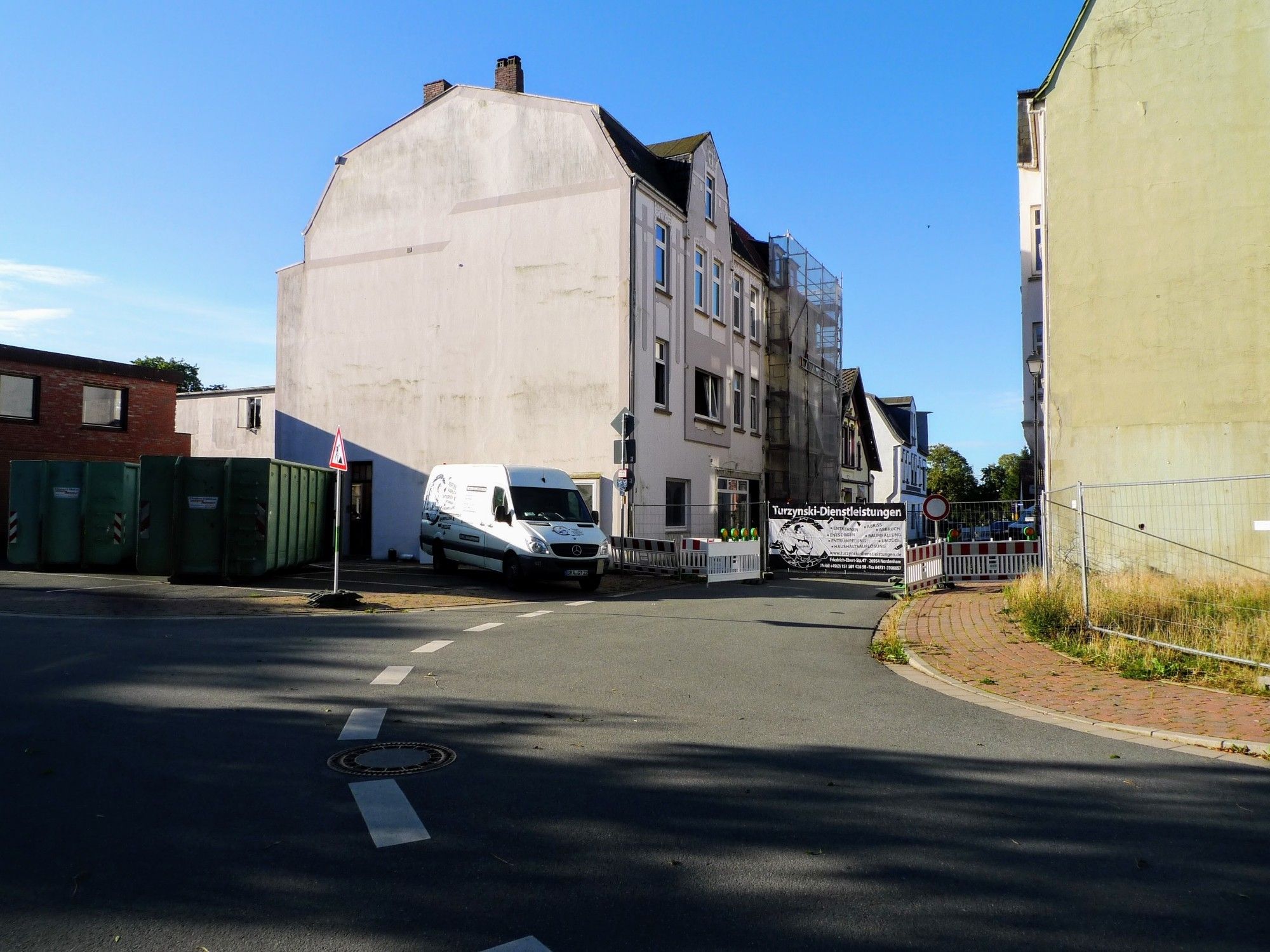 Das Bild zeigt eine städtische Szene mit einer engen Straße, die zwischen mehreren Gebäuden hindurchführt. Auf der linken Seite befinden sich große Abfallcontainer, und ein weißer Transporter steht in der Nähe eines abgesperrten Bereichs. Eines der Gebäude scheint sich im Bau oder in der Renovierung zu befinden, da ein Gerüst angebracht ist. Schilder und Absperrungen deuten darauf hin, dass dieser Abschnitt der Straße für den Verkehr gesperrt ist.