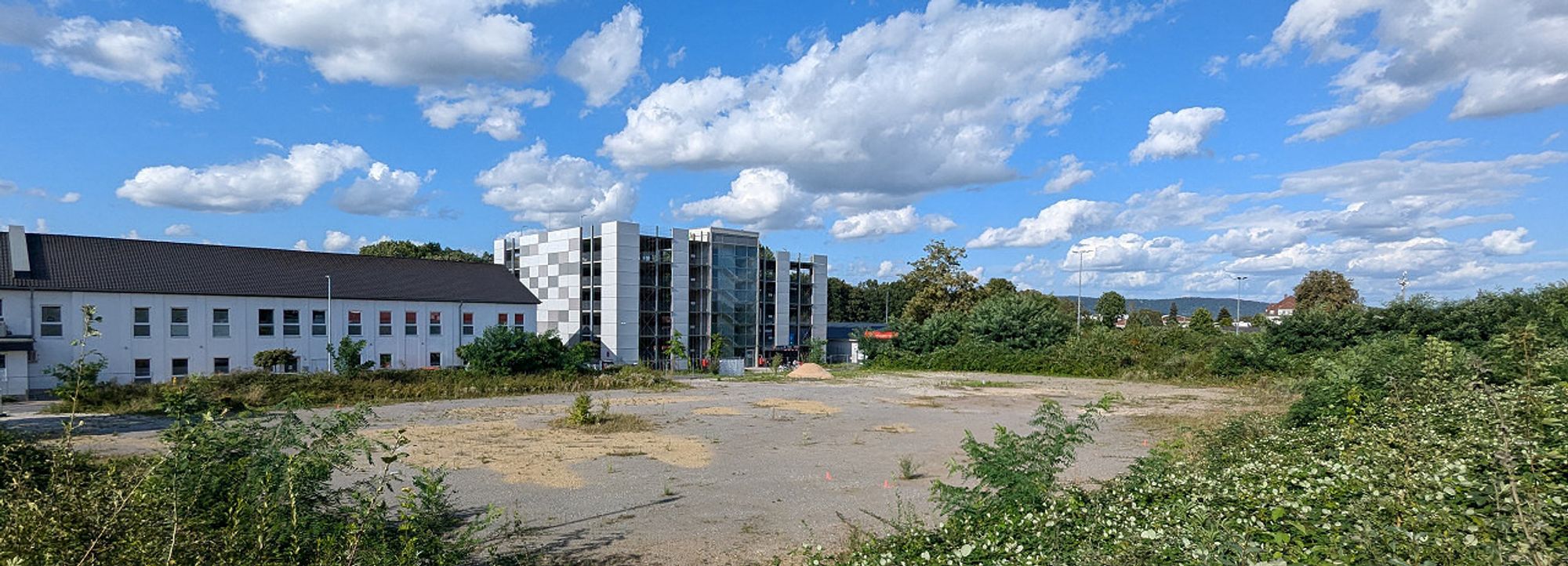 Ein großer, geschotterterte Platz umgeben von einer begrünten Böschung. Abgeschlossen wird er an der hinteren Seite von einem Gebäudekomplex und einem großen Parkhaus.