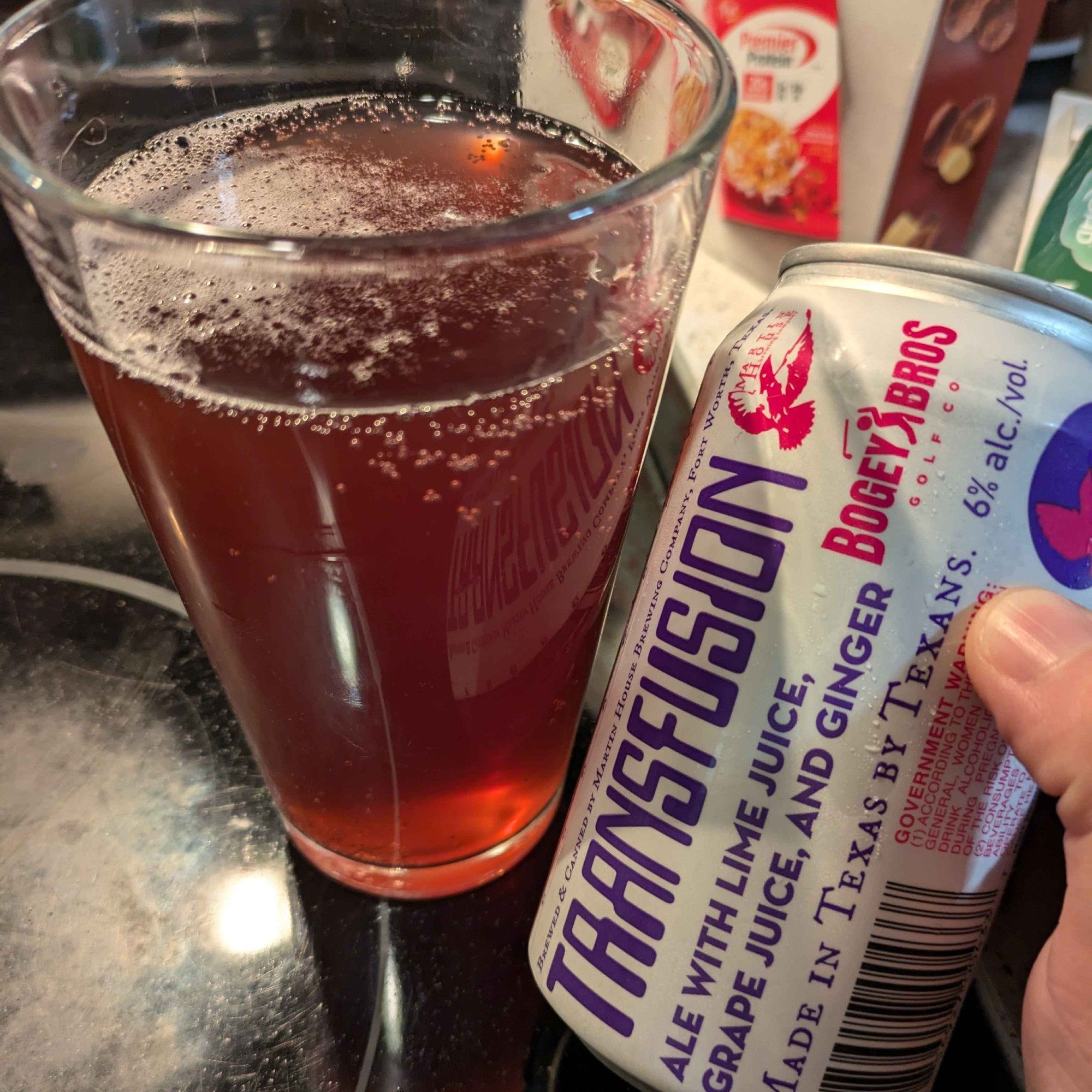 A flat pink drink next to a white and purple can labelled Transfusion Ale with Lime Juice, Grape Juice, and Ginger.