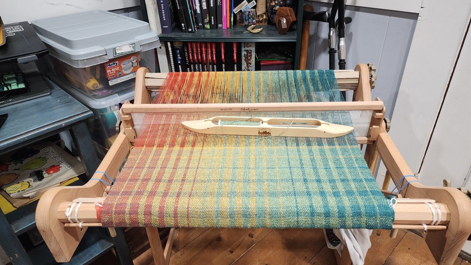 Photo of my rigid heddle loom. The project on it is a wide shawl that uses decreasing and increasing color repeats to transition from orange yarn, to yellow, to green,  to teal in both the warp and weft