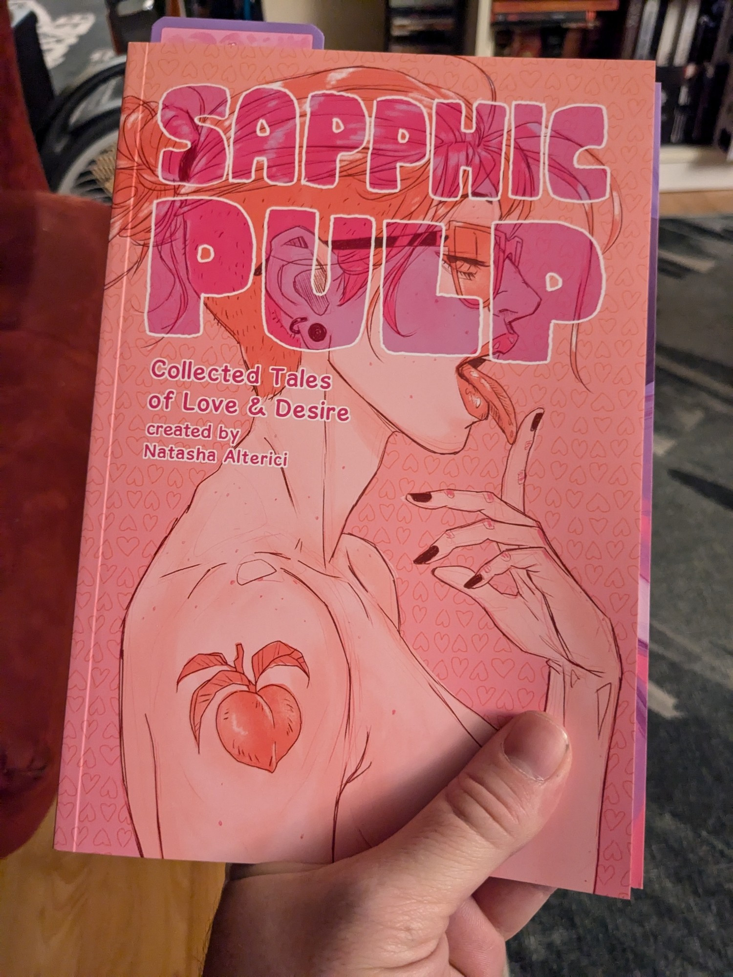 Photo of the erotic anthology, Saphic Pulp, with my thumb cleverly placed to avoid having to put a content warning on my post.