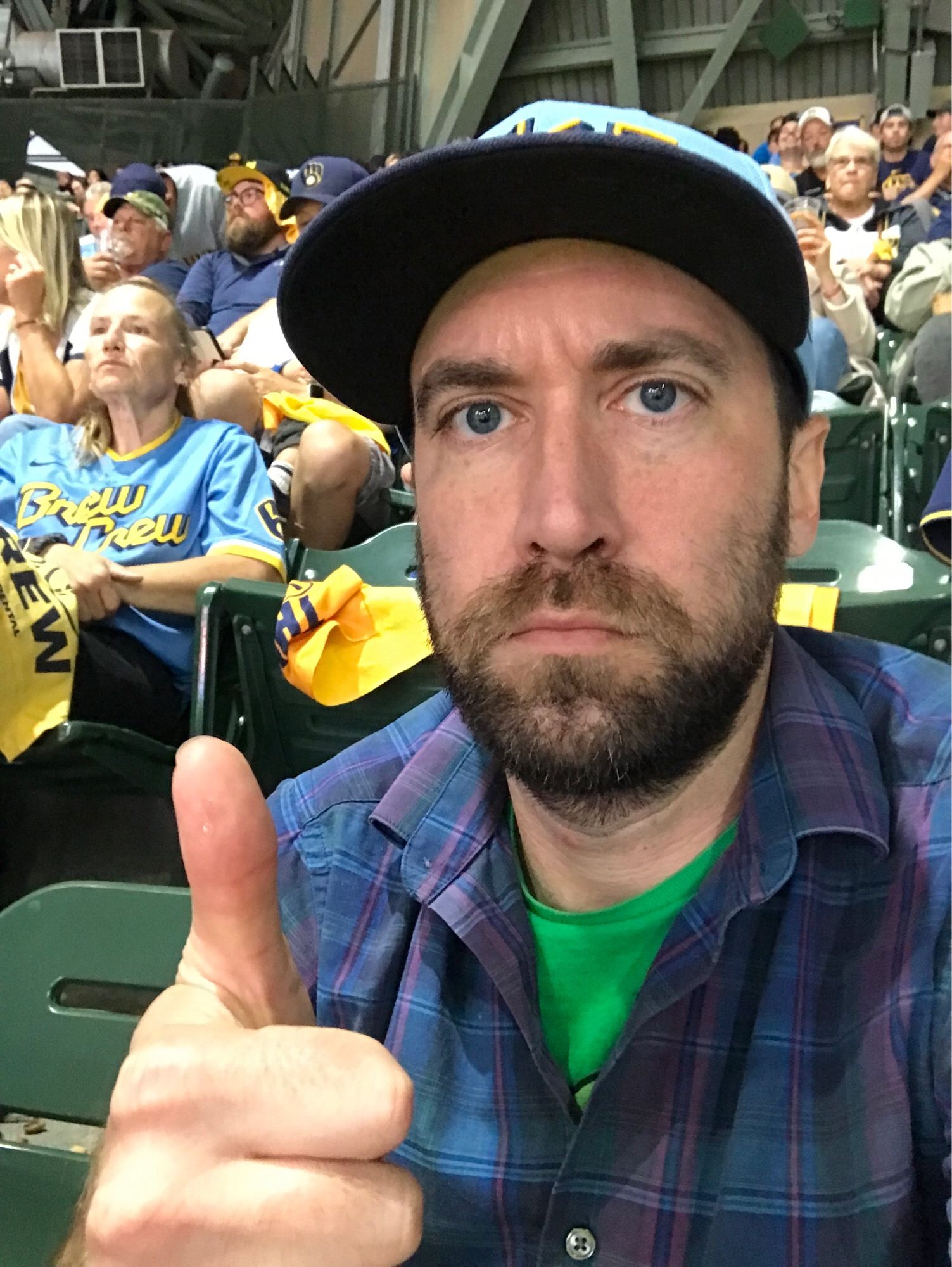 it’s me looking kinda shitty giving a thumbs up surrounded by fellow brewers fans. i’m wearing a blue/purple plaid button down shirt and a green t shirt.