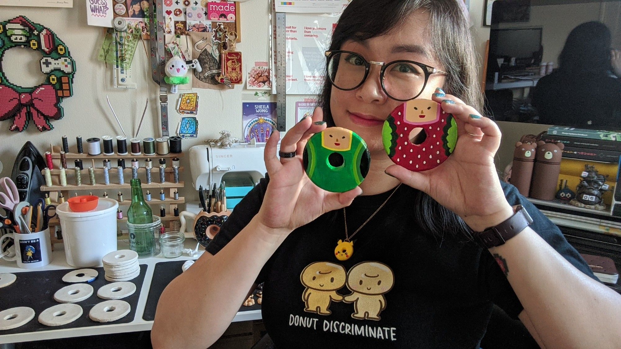 A photo of me, an Asian person with big oversized round glasses smiling while holding up two flat clay donuts painted with Katamari-themed characters (the Prince and Ichigo). In the background is my workspace, including a computer and a bunch of donut-shaped clay on silicone mats, a rack of thread, a mug of scissors, a sewing machine, and just a bunch of art supplies and stuff surrounding me. A happy place.