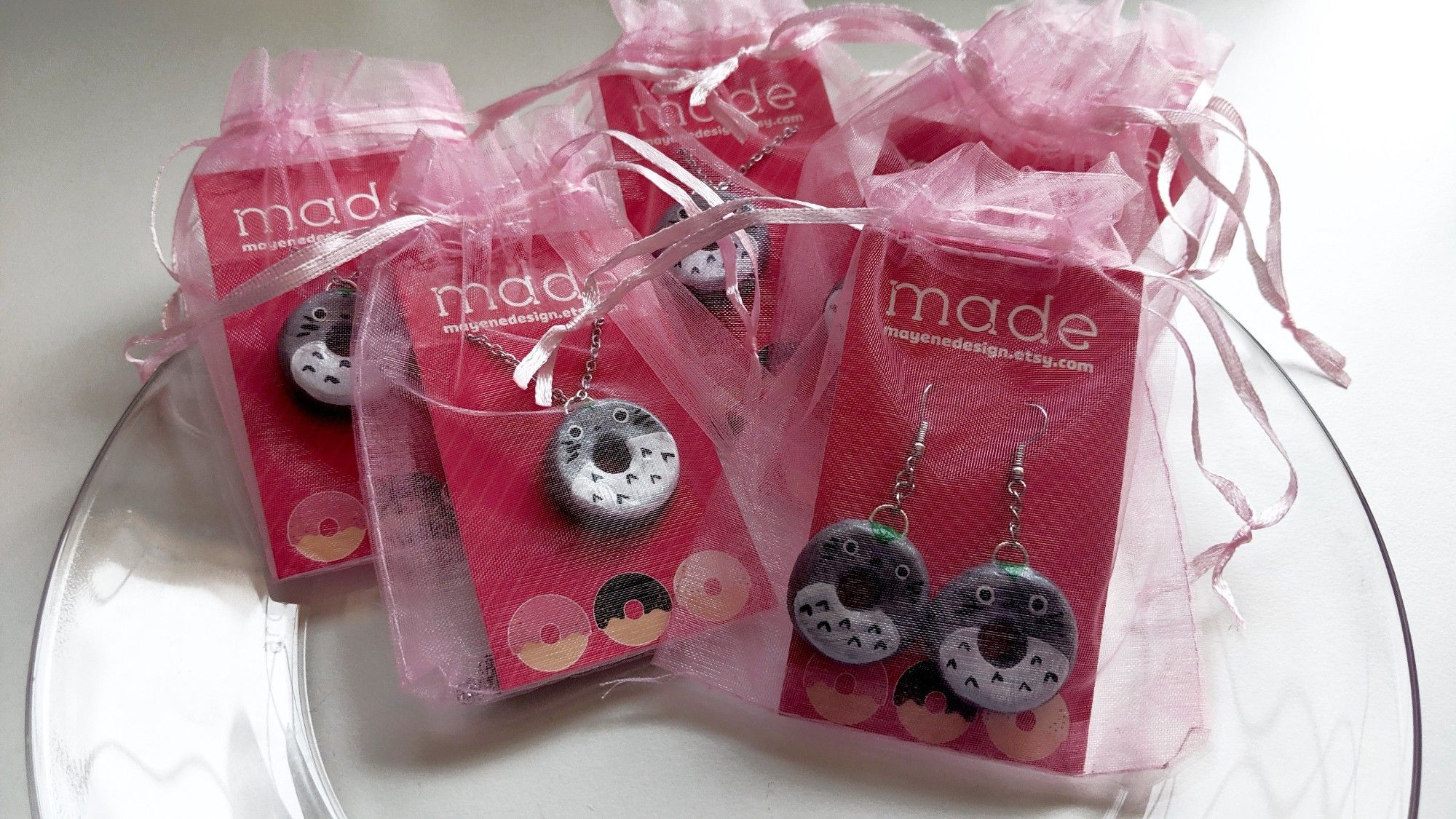 Little packaged up clay donut jewelry, painted like little round Totoros, as necklaces and earring sets on pink cards with my branding on it in pink organza bags laid out on a clear plate.