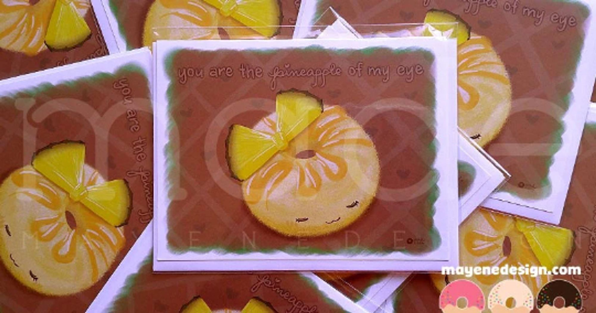 A brown card showing a pineapple donut with a calm face on it, eyes closed, with yellow glaze on top and a couple slices of pineapple topping it. The card says: "You are the pineapple of my eye."