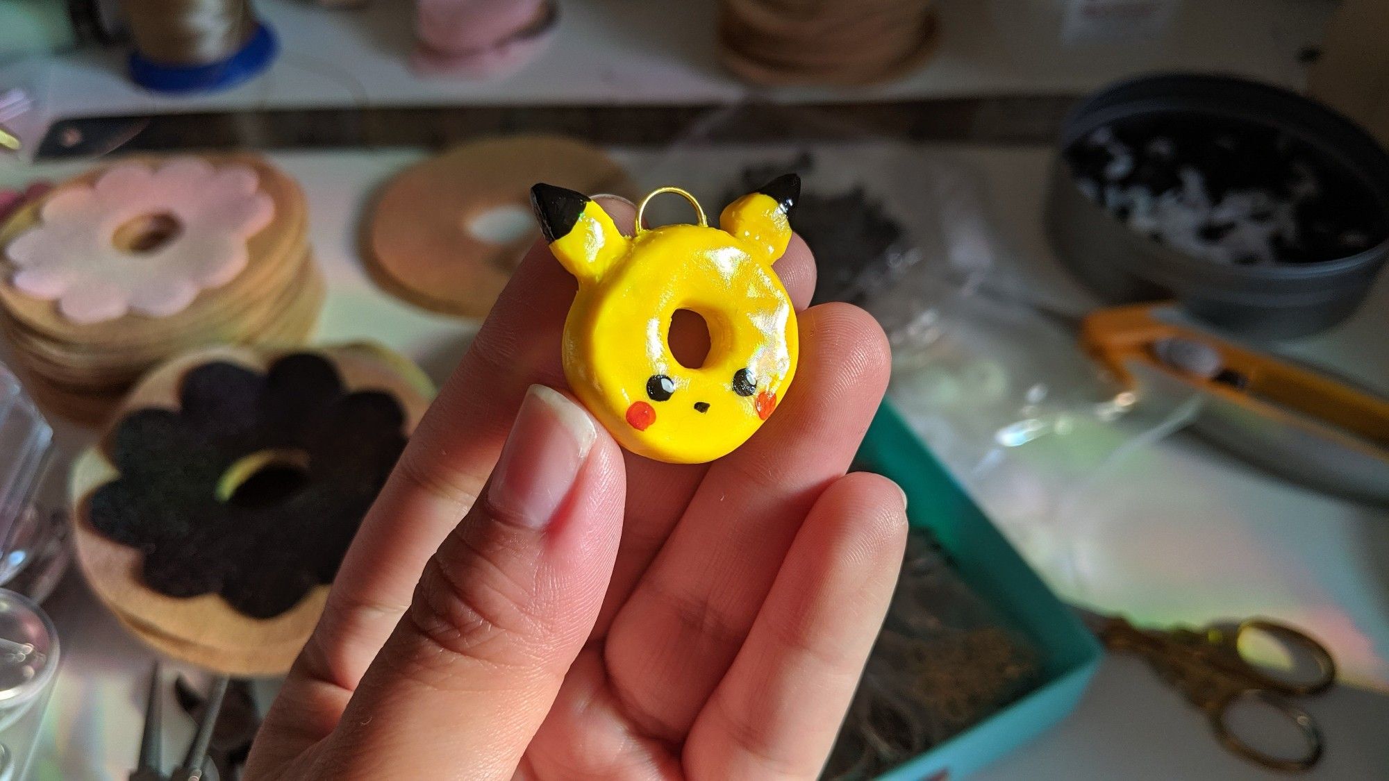 A finished clay pendant, glossy and painted mostly yellow. A small donut-shaped pendant with ears sticking out themed after Pikachu, held in my hand. In the background, felt donut-themed crafts and scissors are in the background.