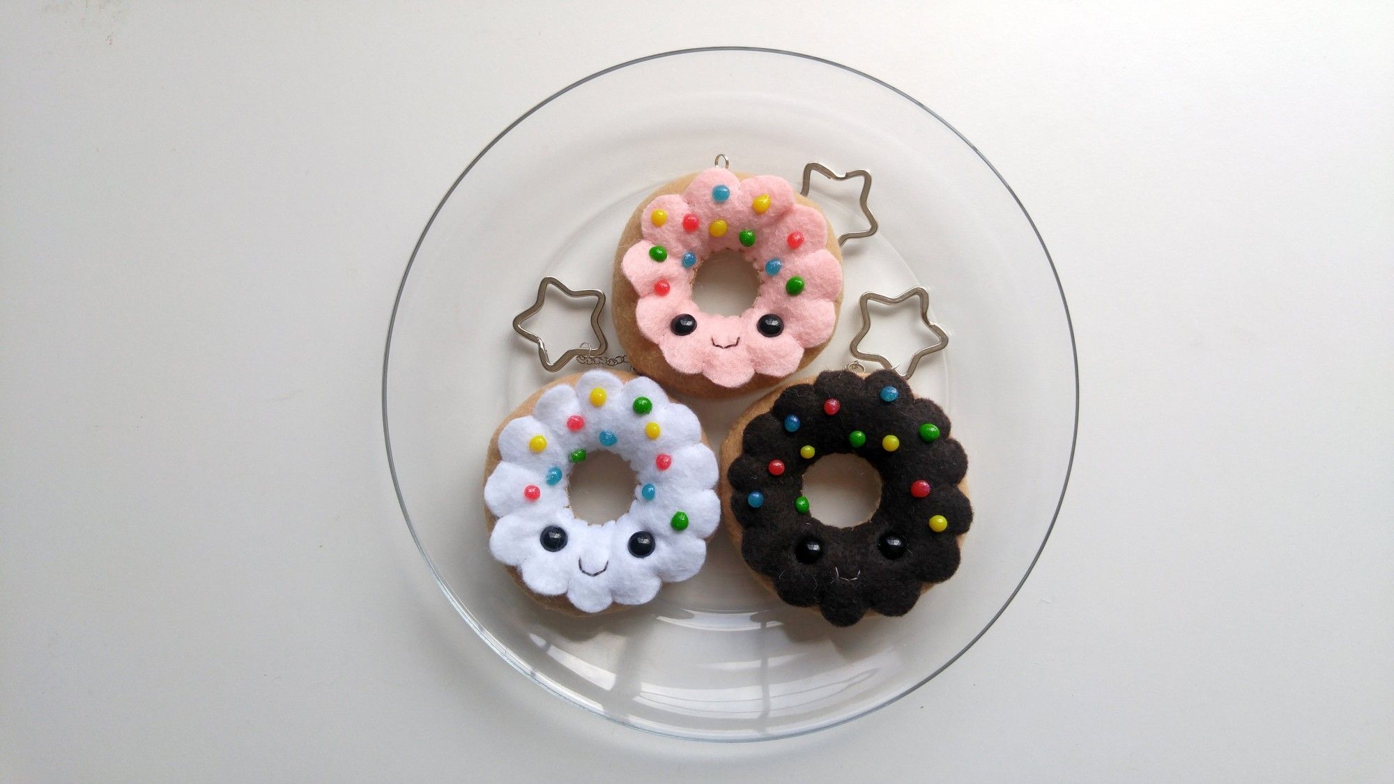 Top down view of a clear plate on a white desk with three handmade felt donut keychains, each with a cute little smiley face and sprinkles on top. The attached keychains are in the shape of stars.