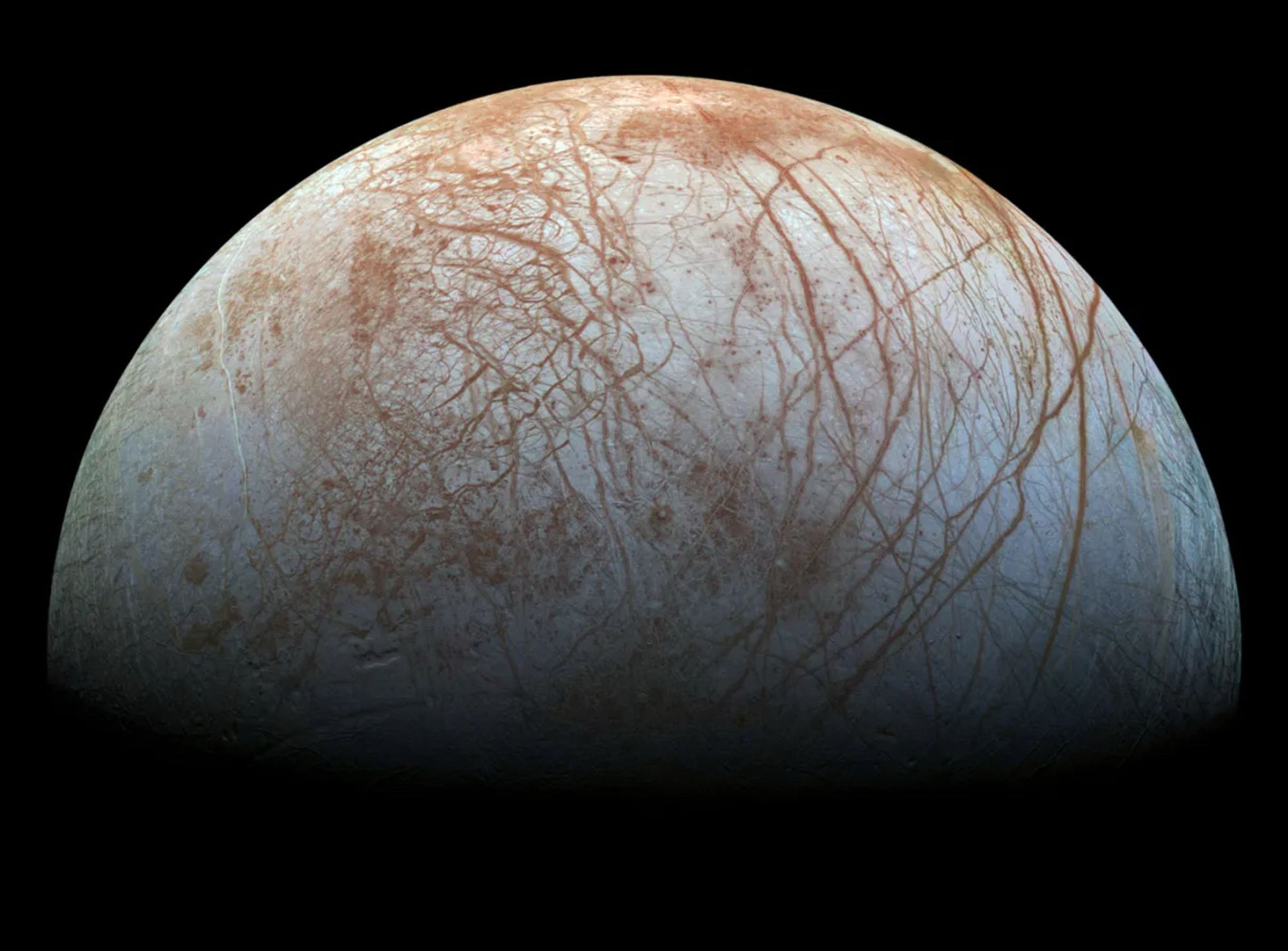 A close to true colour image of  Jupiter’s moon, Europa, showing long reddish cracks running through its lightly coloured grey- blue icy surface.