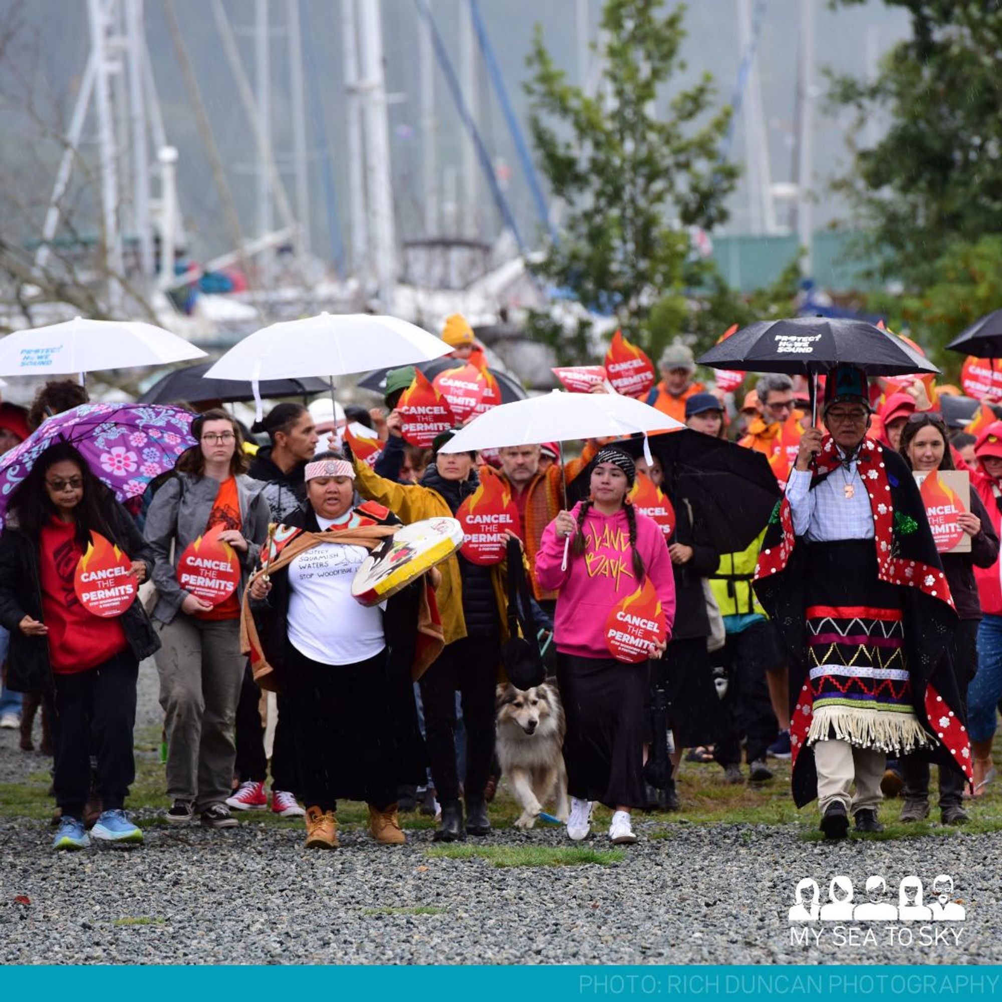 Indigenous leaders and settlers march carrying umbrellas and signs in the shape of a fire emoji that read "Cancel the Permits."