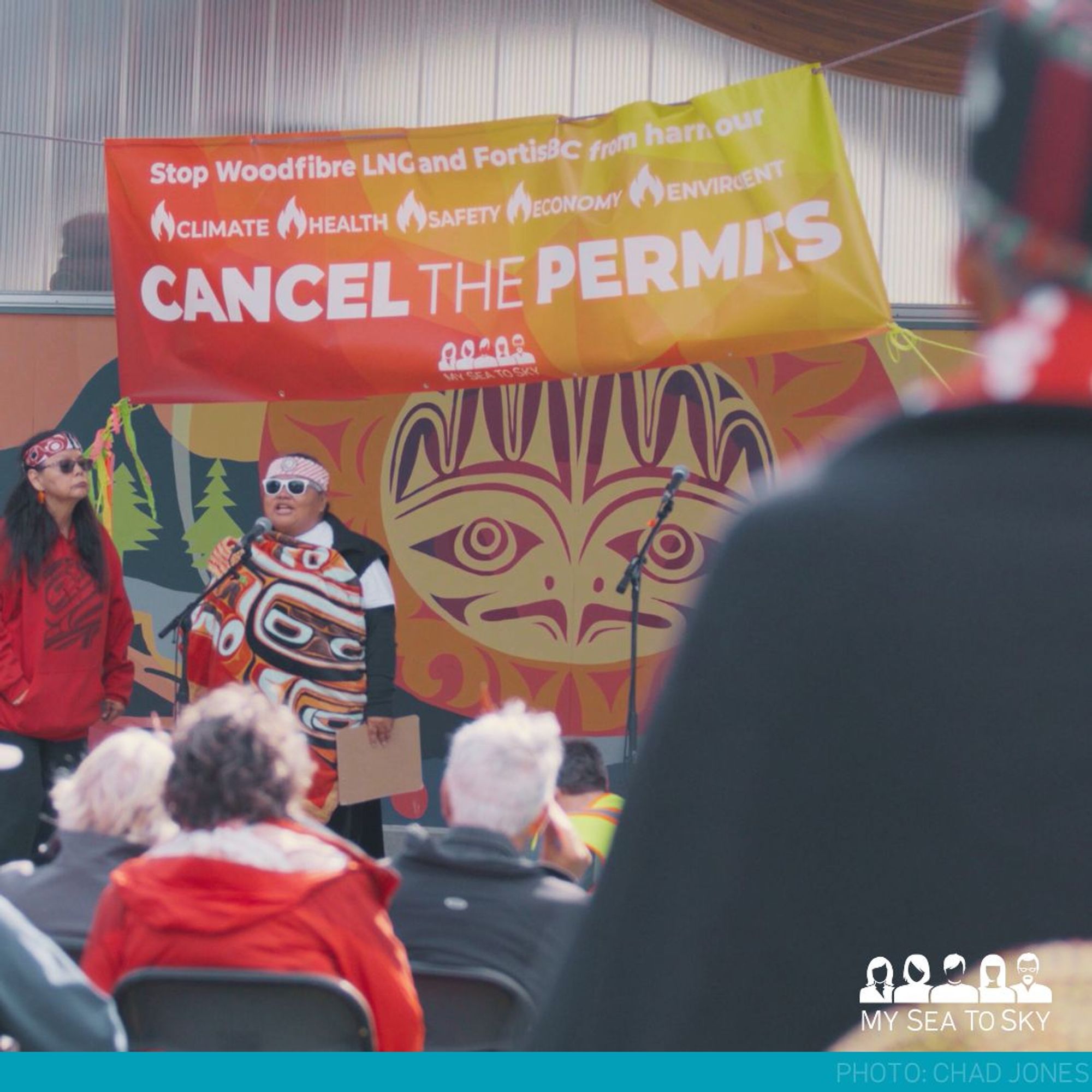 Chief Na'Moks listens to Squamish Nation Elder Tioutenaat Jackie Williams and her daughter Sewainchelwat Jaime Williams for a Witness Ceremony. A red, orange, and yellow banner reads "Stop Woodfibre LNG and FortisBC from harming our Climate Health Safety Economy Environment. Cancel the Permits."