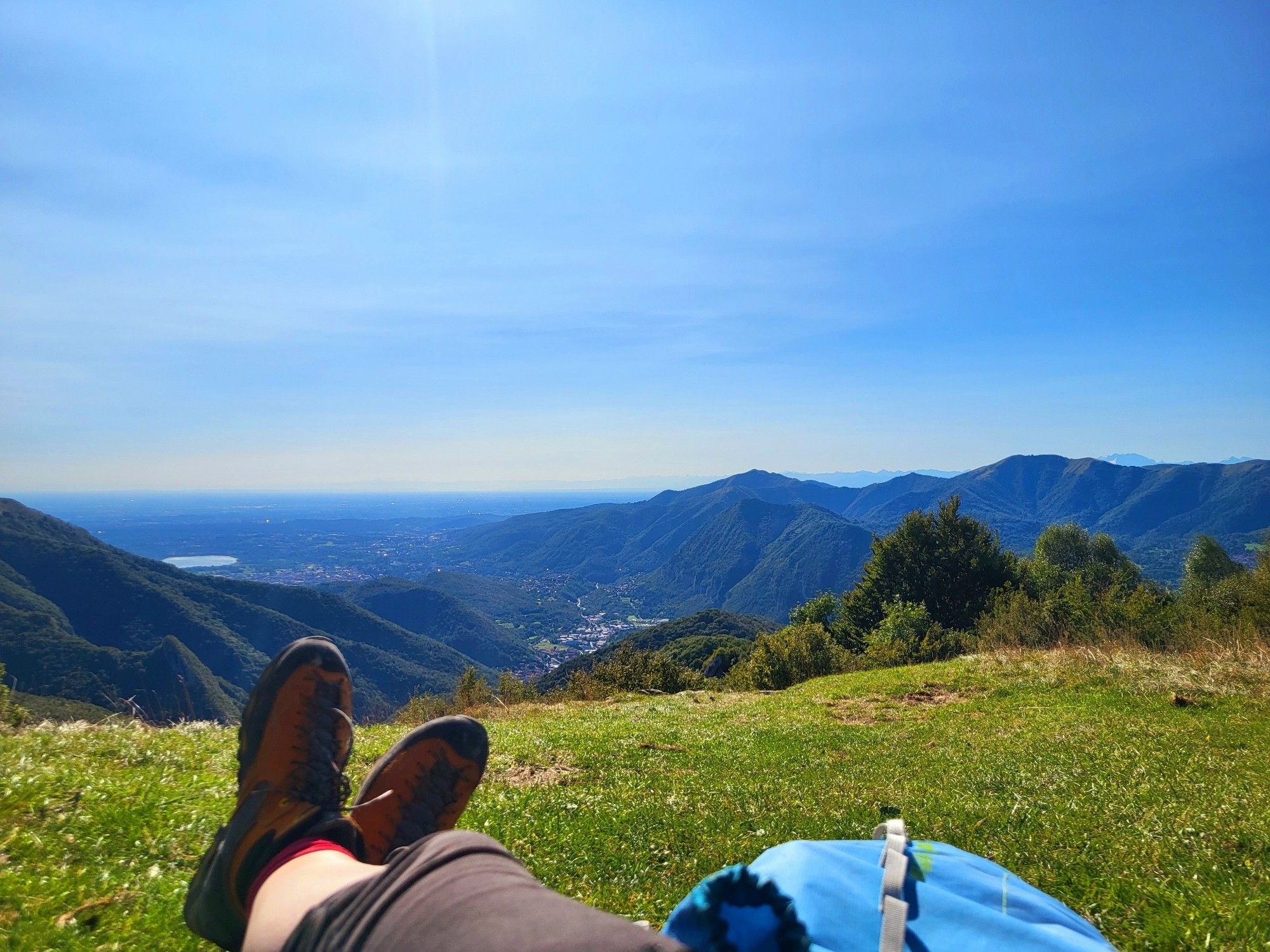 Rast auf einer Bergwiese, im Bild meine Beine, Bergstiefel, der Rucksack, im Hintergrund der Fernblick aif dwn südlichen Alpenrand