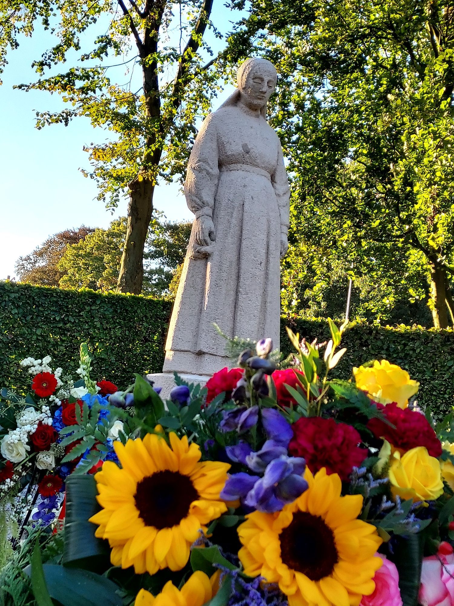 Krans met verschillende bloemen waaronder twee opvallende zonnebloemen. Op de achtergrond staat het standbeeld van het vrouwtje van Putten.