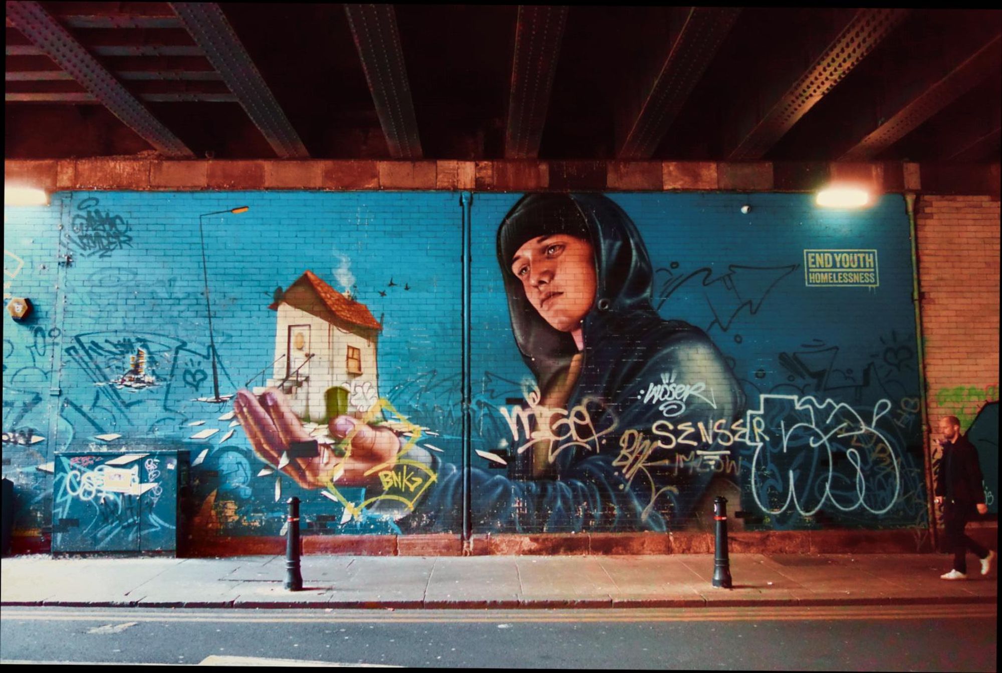 A 16:9 ratio piece of photo realistic street mural artwork under a railway bridge. A stencil type logo of white e text on green proclaims "end youth homelessness" on the top right.
An aquasmarine graduated blue background backs a young white man with dark hair in a hoodie with his aeft arm outstretched. In his palm is a small idealised house with water droplets falling from it.
There is unrerlated basic graffiti around the image.