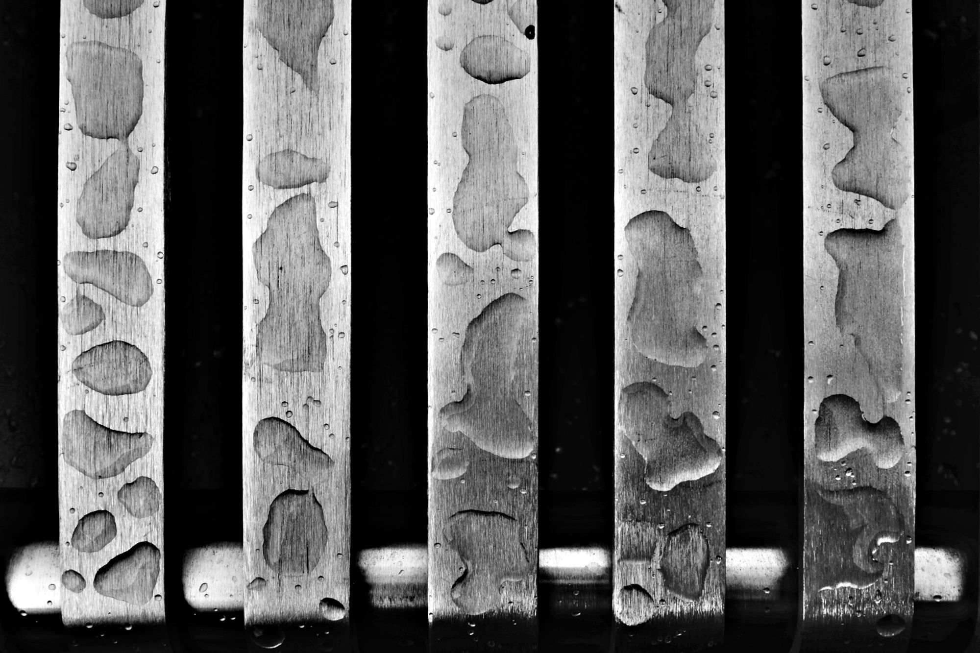 A high contrast photo of a stanless steel bench. The silver veertical slatts of the bench are dotted with large rain droplets. The gap inbetween is pure black. The structure of the metal can been seen in detail whilst the water puddles magnidy the metal structure in parts.