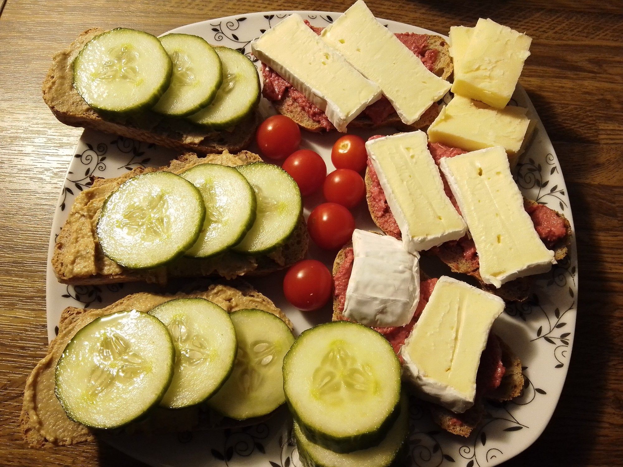 Foto eines weißen Tellers mit schwarzen Rankenornamenten am Rand der auf einer dunkelbraunen Küchenarbeitsplatte steht. Der Blickwinkel ist von oben auf den Teller. Zu sehen sind 6 Scheiben belegtes Baguette dazwischen kleine Tomaten, Gurkenscheiben und drei Stücke heller Cheddar Käse. Das Baguette ist ein dunkles mit Walnuss und Feige. Drei Scheiben sind mit der veganen Streichwurst von Rossmann und Gurkenscheiben belegt. Die anderen drei mit einem rote Beete Meerrettich Aufstrich, ebenfalls von Rossmann, und Camembert Streifen.