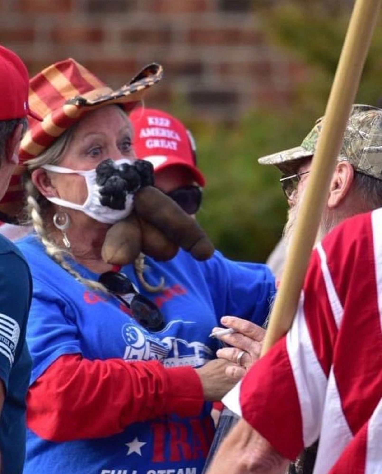 Foto einer MAGA Frau auf, höchstwahrscheinlich, einer Trump Ralley. Sie trägt die obligatorischen Patriot Farben blau rot weiß sowie ein blaues Shirt mit dem aktuellen Wahlkampf Logo über ihrem roten Pullover. Dazu ein Cowboy Hut. Das wirklich furchtbare an diesem Bild ist aber ihre selbst gebastelte Verballhornung einer ffp2 Maske. Es sieht aus wie ein weißer Tanga aus dem ein riesiger schwarzer Penis und Hoden mit Schamhaar heraus schauen.