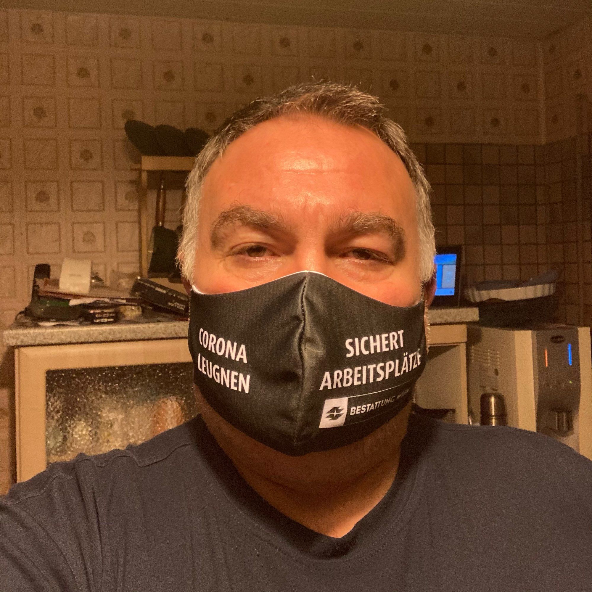 Mann in schwarzem T-Shirt und einer Stoffmaske mit der Aufschrift „Corona leugnen sichert Arbeitsplätze“ von der Firma Bestattung Wien, aufgenommen bei künstlichem Licht in einer Küche.