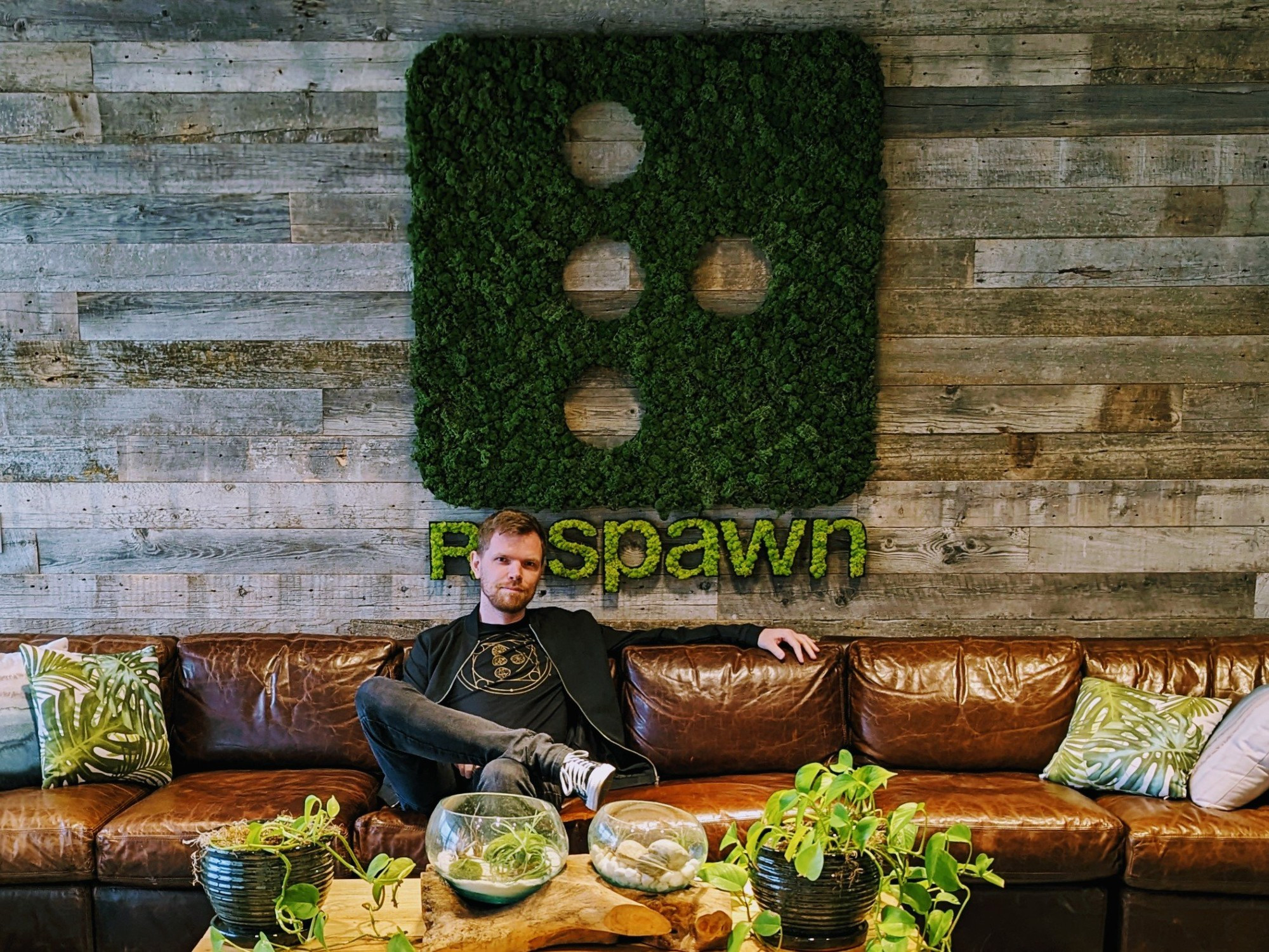 Me at the Respawn office, sitting on a brown leather couch with several green and grey cushions. Behind me is a wall made of a mix of grey and brown wooden planks, with a large Respawn logo made of tight green foliage. In front of me sits a coffee table with various potted plants.