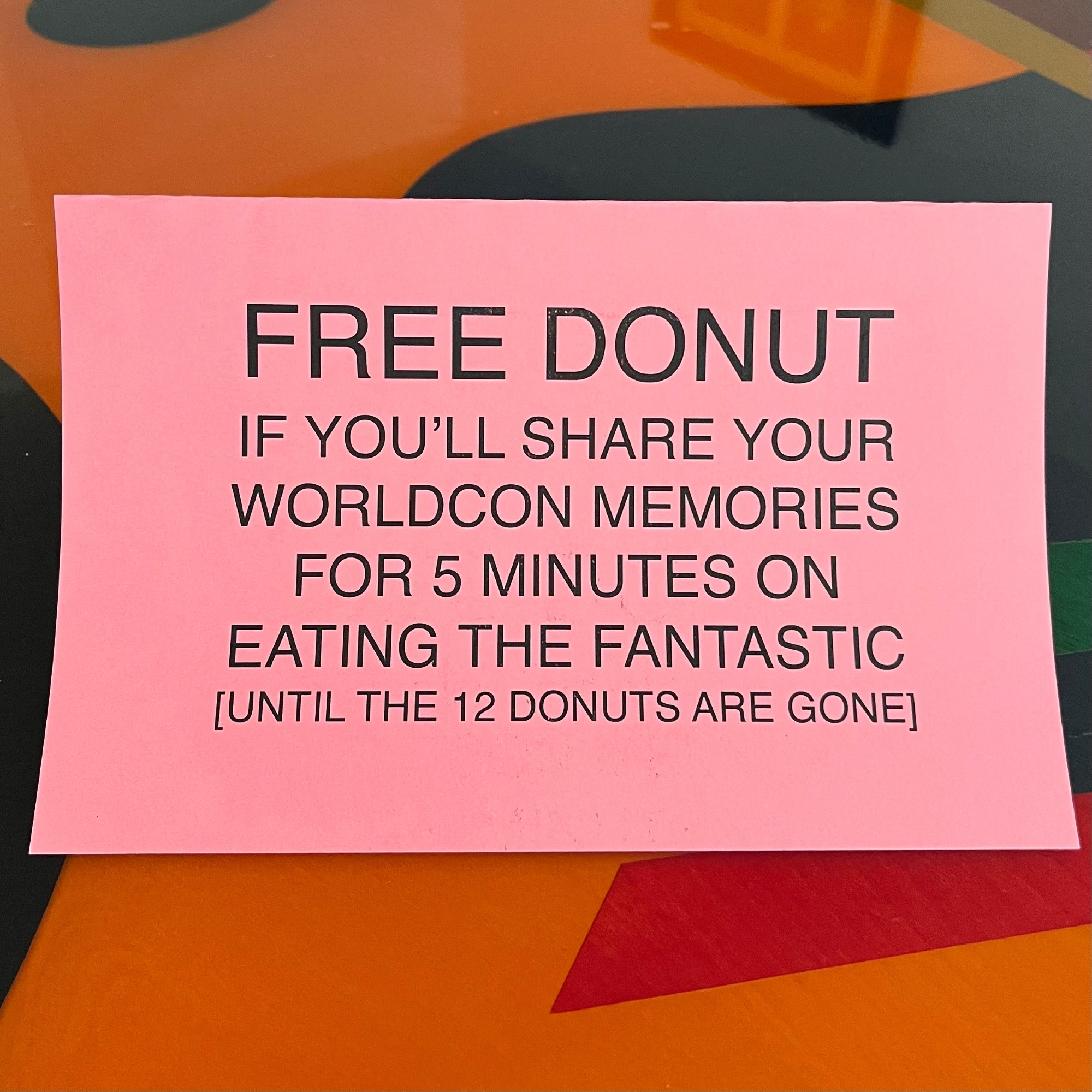 Sign in black ink on pink paper. It's a multi-colored table. The sign reads:

FREE DONUT
IF YOU’LL SHARE YOUR WORLDCON MEMORIES  FOR 5 MINUTES ON  EATING THE FANTASTIC
[UNTIL THE 12 DONUTS ARE GONE]