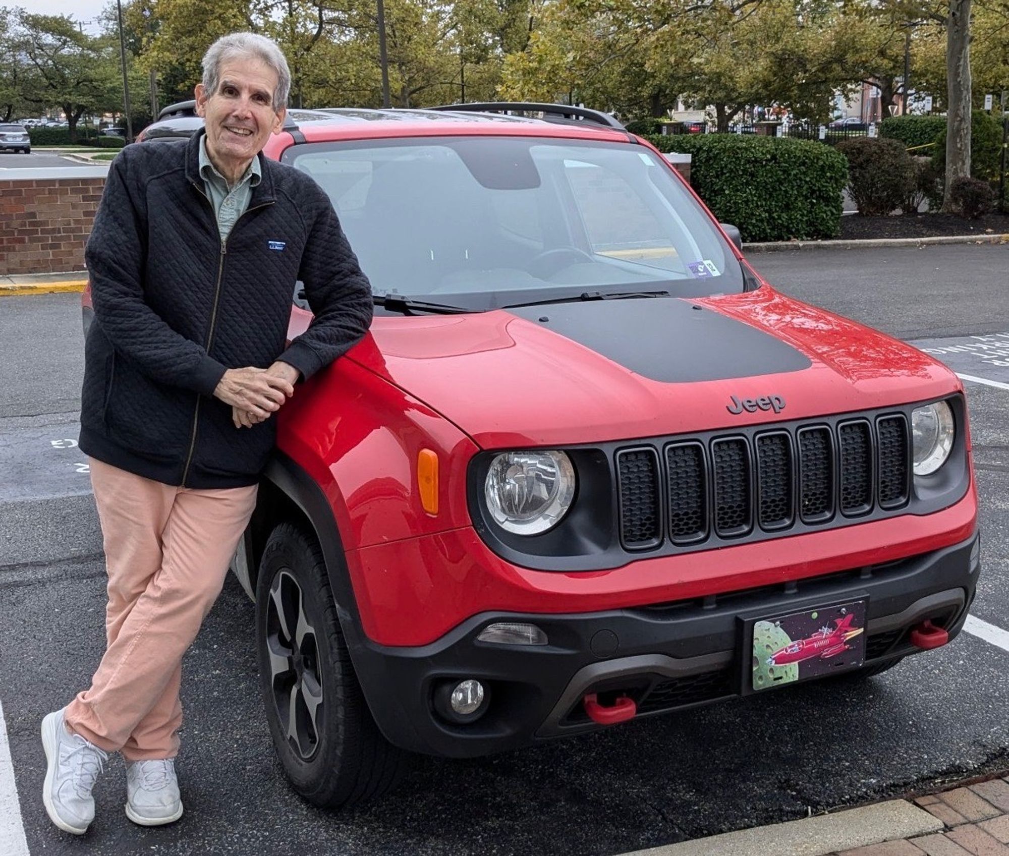 Me, wearing a black zippered sweatshirt and salmon-colored pants, leaning against my bright red 2019 Jeep Renegade Trailhawk. West Virginia doesn't require front license plates, so in that spot is metal plaque of the same shape depicting a red rocket ship that might have appeared on a 1940's science fiction magazine.